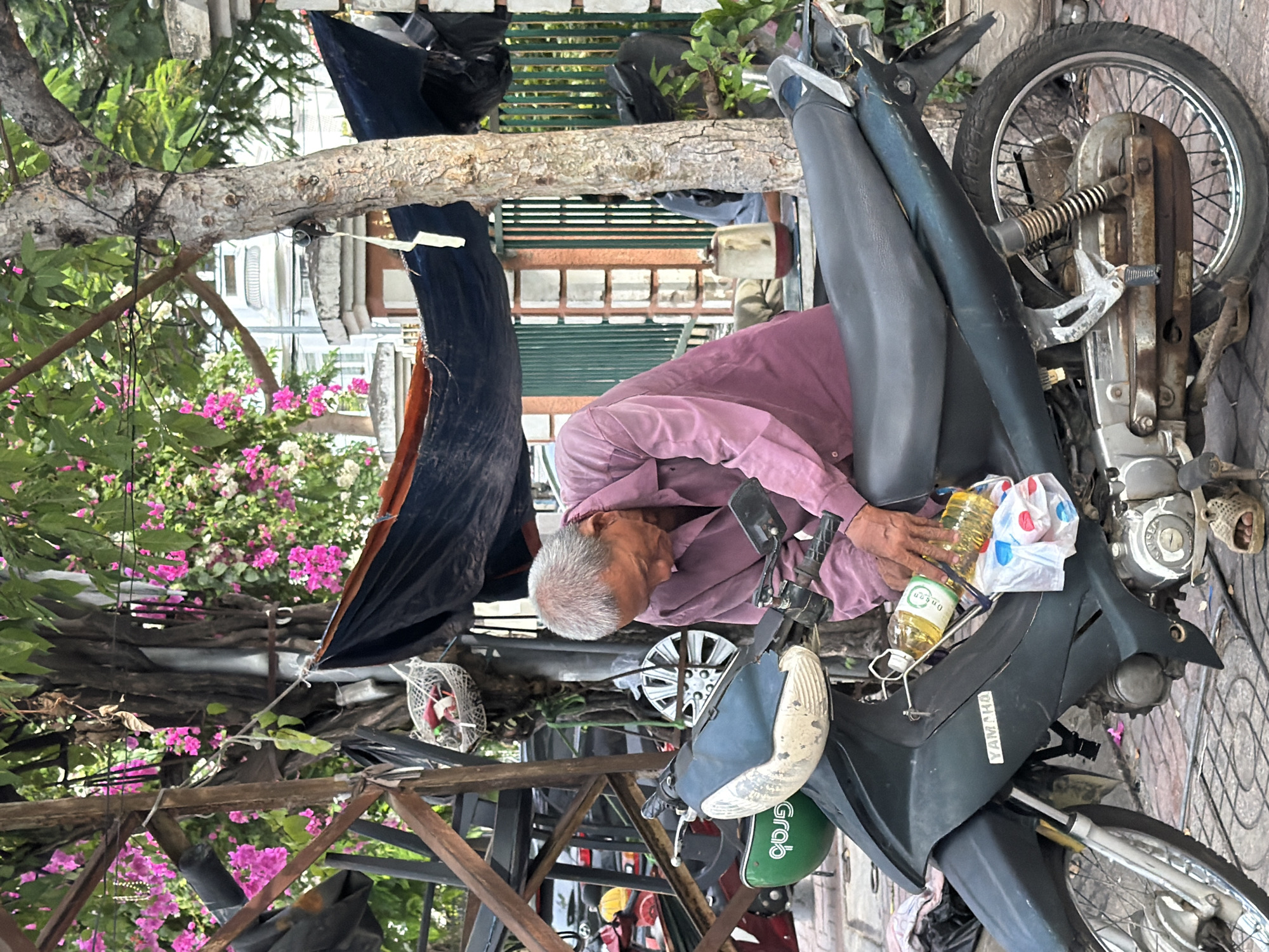 An elderly man with graying hair, wearing a purple long-sleeved shirt, is crouched next to a dark green motorcycle. He is holding two bottles of cooking oil, one in each hand, and appears to be placing them in or retrieving them from a holder on the motorcycle. The motorcycle is parked on a paved surface, likely a sidewalk, in what appears to be a busy urban environment. Behind the man and the motorcycle, a large tree with pink and white flowers is visible, along with some makeshift structures covered with tarps. A green helmet with the word "GRAB" on it is attached to the front of the motorcycle. In the background, there are buildings and a green metal fence. The overall scene suggests a typical street-side setting in Vietnam. The presence of the Grab helmet indicates the potential use of the motorcycle for ride-sharing services. The cluttered environment with tarps and makeshift structures suggests a location that may be used for commerce or temporary housing.