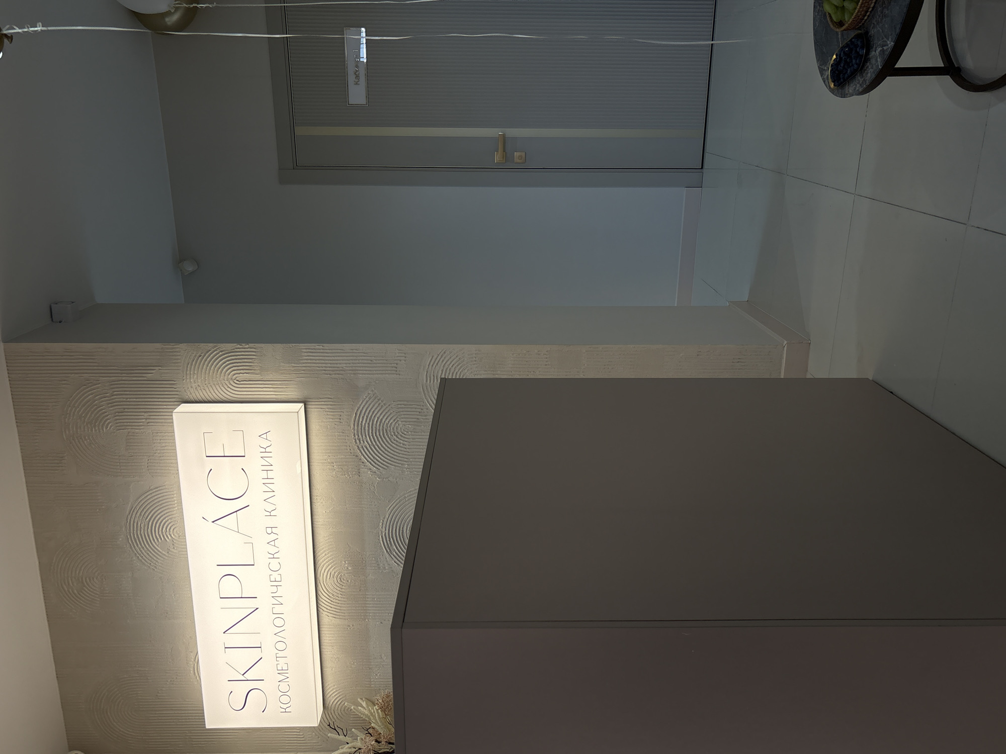 An interior view of a modern reception area within a beauty clinic, identified as "SKINPLACE КОСМЕТОЛОГИЧЕСКАЯ КЛИНИКА" (SKINPLACE COSMETOLOGICAL CLINIC), located in Rostov-on-Don, Russia. The reception desk, a large, dark gray, rectangular unit, occupies the left foreground. Behind it, a wall features textured decorative panels and a backlit sign bearing the clinic's name in English and Russian. To the right, a closed door with a vertical textured design and a golden handle is visible. A small sign above the door reads "Кабинет 1" (Office 1). In the lower right corner, a small, dark-colored coffee table with a marble-patterned top holds a basket of green fruit and a dark oval dish, possibly containing berries. The floor is tiled with light-colored square tiles. The overall ambiance is clean, minimalist, and professional.
