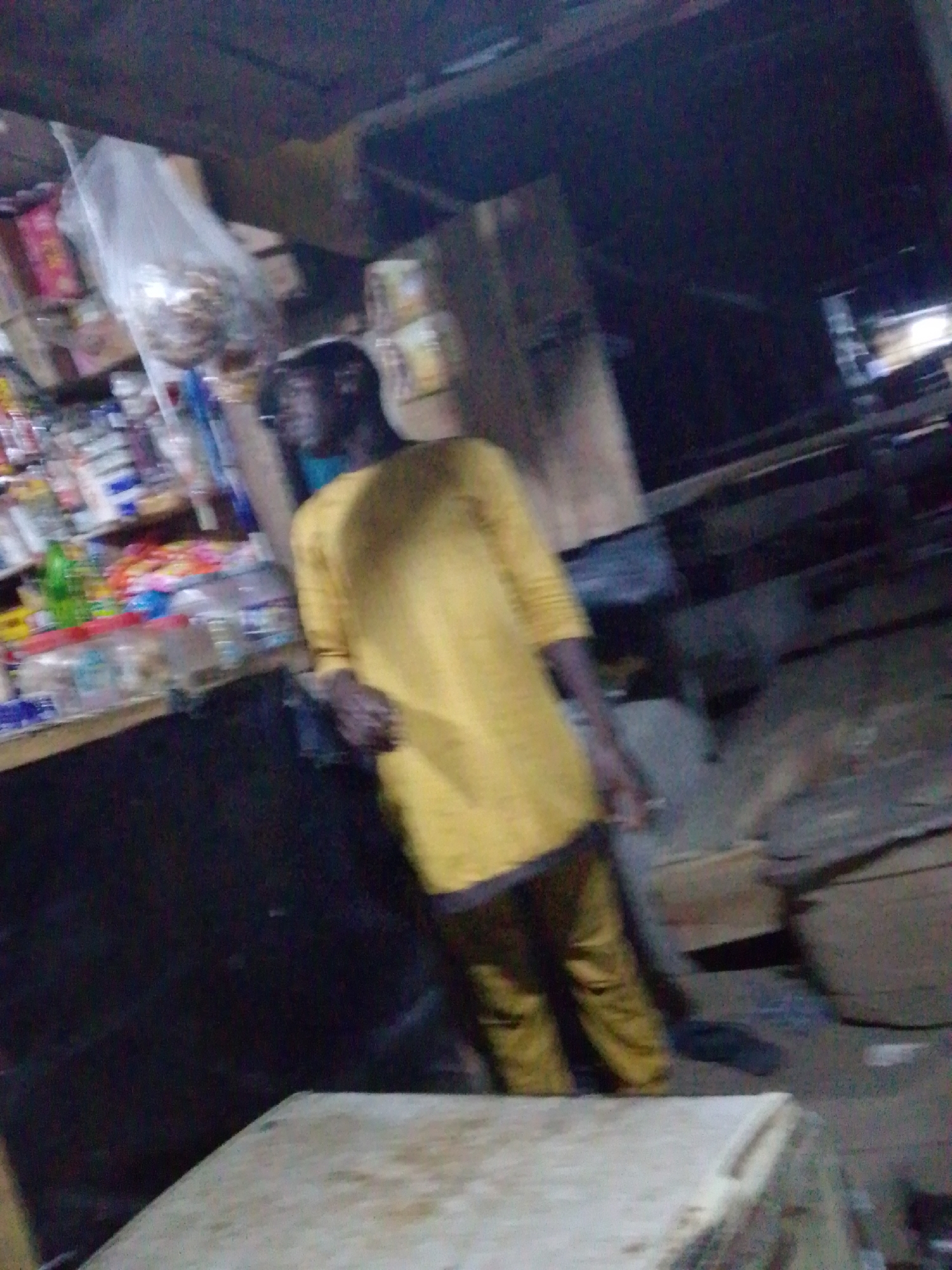 The image shows a dimly lit shop in Ajaokuta, Nigeria. A man in a yellow long-sleeved shirt and matching pants stands leaning against a dark counter. The shop has shelves stocked with various items, including food products in plastic packaging. The surrounding environment appears cluttered with miscellaneous objects.