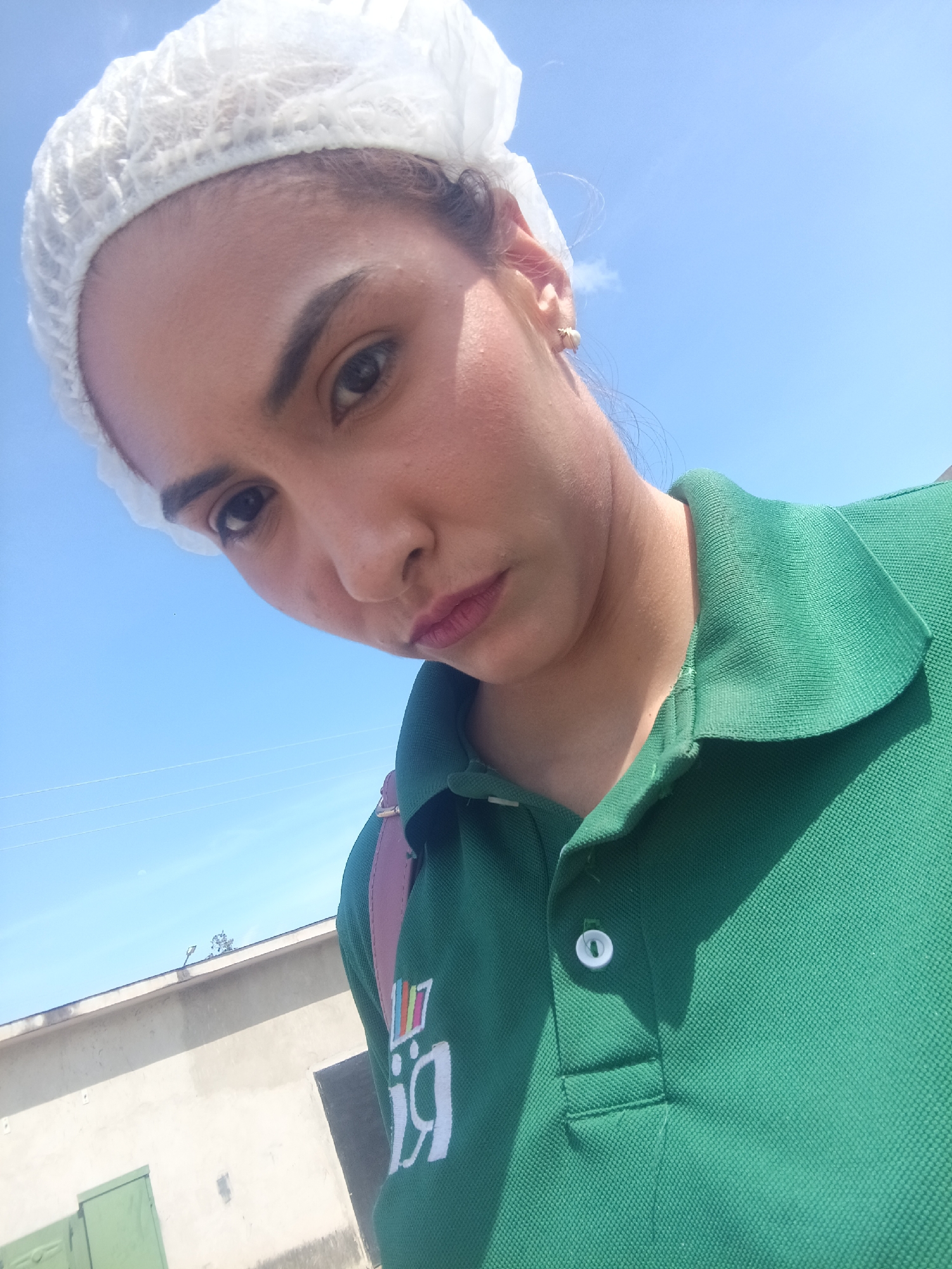 A medium shot captures a woman wearing a white hairnet and a green polo shirt with an inverted logo. The sky is bright blue with a few wispy clouds. A building with a grey facade, green doors, and a dark window is visible in the background. The woman's gaze is directed down, and she is positioned at a high angle, looking down at the camera. This scene is located in Juan Griego, Venezuela.