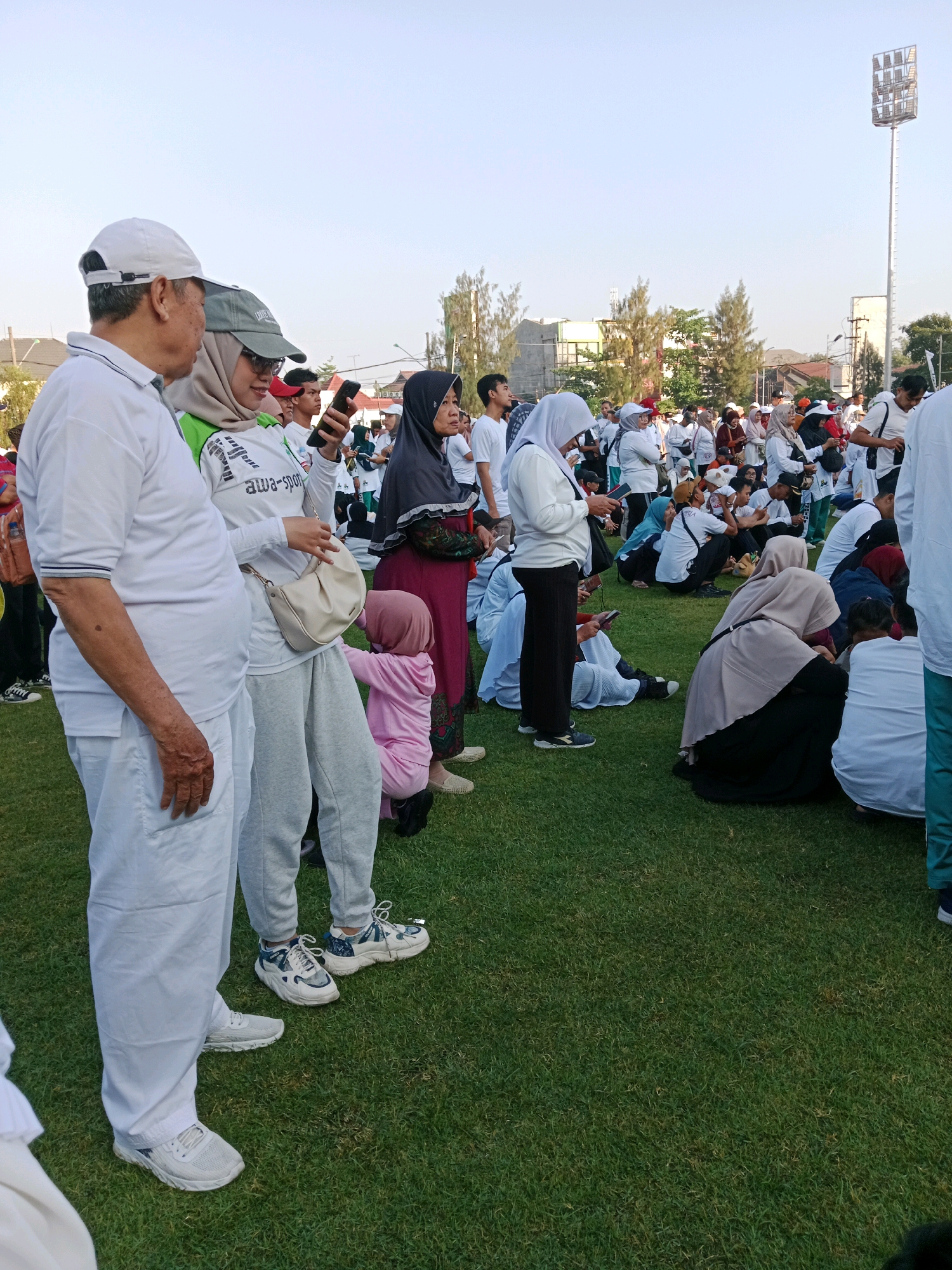 The image depicts a large gathering of people outdoors on a grassy field, likely in the city of Surakarta, Indonesia, under a clear sky. In the foreground, an elderly man in a white polo shirt and white trousers stands to the left, looking towards a woman next to him. The woman, wearing a light-colored hijab and a green and white shirt with the text "awa-spor", holds a smartphone in her hand and appears to be looking at it or showing something on it to the man. She is also wearing light grey athletic pants and carrying a small cream-colored shoulder bag. To the right of this pair, a young child in a pink outfit is partially visible, interacting with the woman.
The background reveals a dense crowd of people, many wearing white attire and headscarves, suggesting a communal event or activity. Some individuals are seated on the grass, while others are standing. Buildings and trees are visible in the mid-ground, and a prominent stadium light tower stands tall in the distance on the right. The overall scene suggests a daytime event, possibly a sporting activity, a religious gathering, or a community festival. The presence of the stadium light tower indicates a potential location near a sports facility.