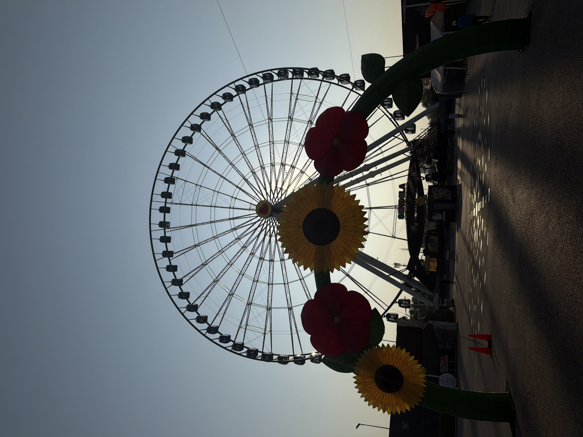 The image depicts the "Estrella de Puebla," a Ferris wheel in Puebla, Mexico, against a pale sky. The wheel's structure is visible, with passenger cars attached. In the foreground, there are decorative artificial flowers – two sunflowers and two red flowers – attached to green stems. The ground is paved and dark, suggesting possible wetness. The image is taken from a low angle, looking upward at the wheel and the sky.