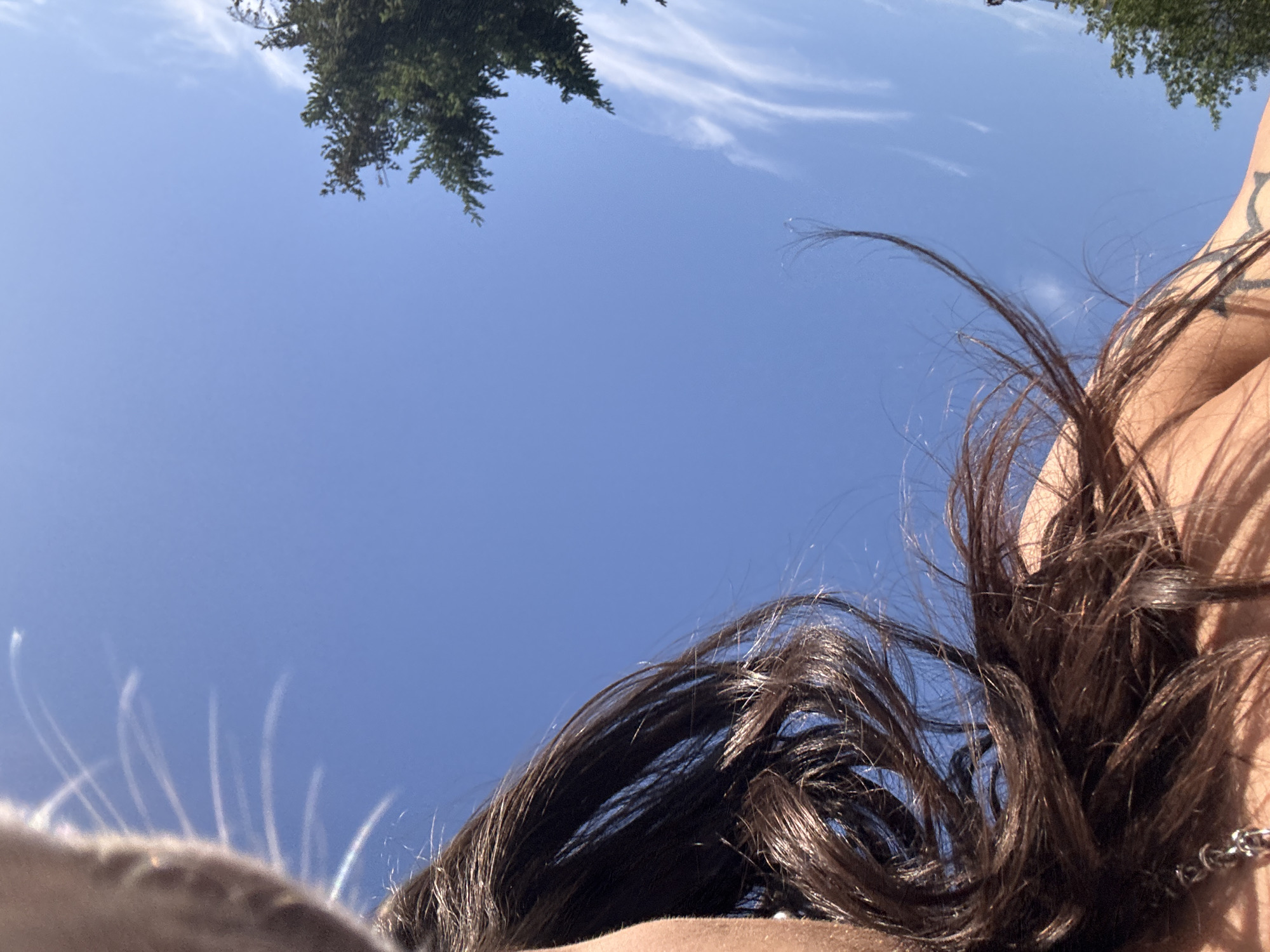 The image is taken from a low angle, looking upwards towards a bright blue sky. A cluster of green tree branches is visible in the top left corner. On the right side of the frame, there is a portion of a person's shoulder, neck, and hair. The hair is dark brown and disheveled, with strands extending across the lower part of the image. A silver chain necklace is visible around the neck. There are no other discernible objects or people in the frame. The location is not identifiable as Sofia, Bulgaria, from this image alone.