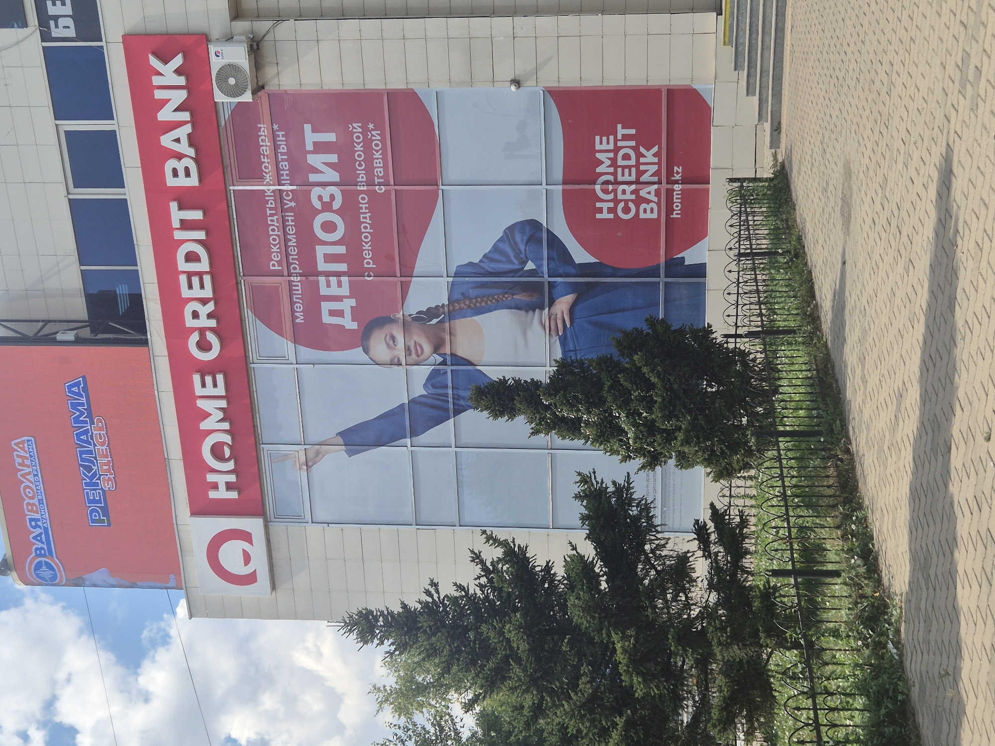 The image shows the facade of a Home Credit Bank branch in Kokshetau, Kazakhstan. The building has a modern, light-colored facade with large glass windows. A prominent advertisement for Home Credit Bank is displayed on the building, featuring a young woman in a blue suit and the text "HOME CREDIT BANK" and "ДЕПОЗИТ". The advertisement is in Russian and Kazakh. To the left of the bank's entrance, there are several green coniferous trees. A metal fence separates the sidewalk from the building. The sidewalk is paved with light-colored interlocking stones. The sky is blue with scattered white clouds.