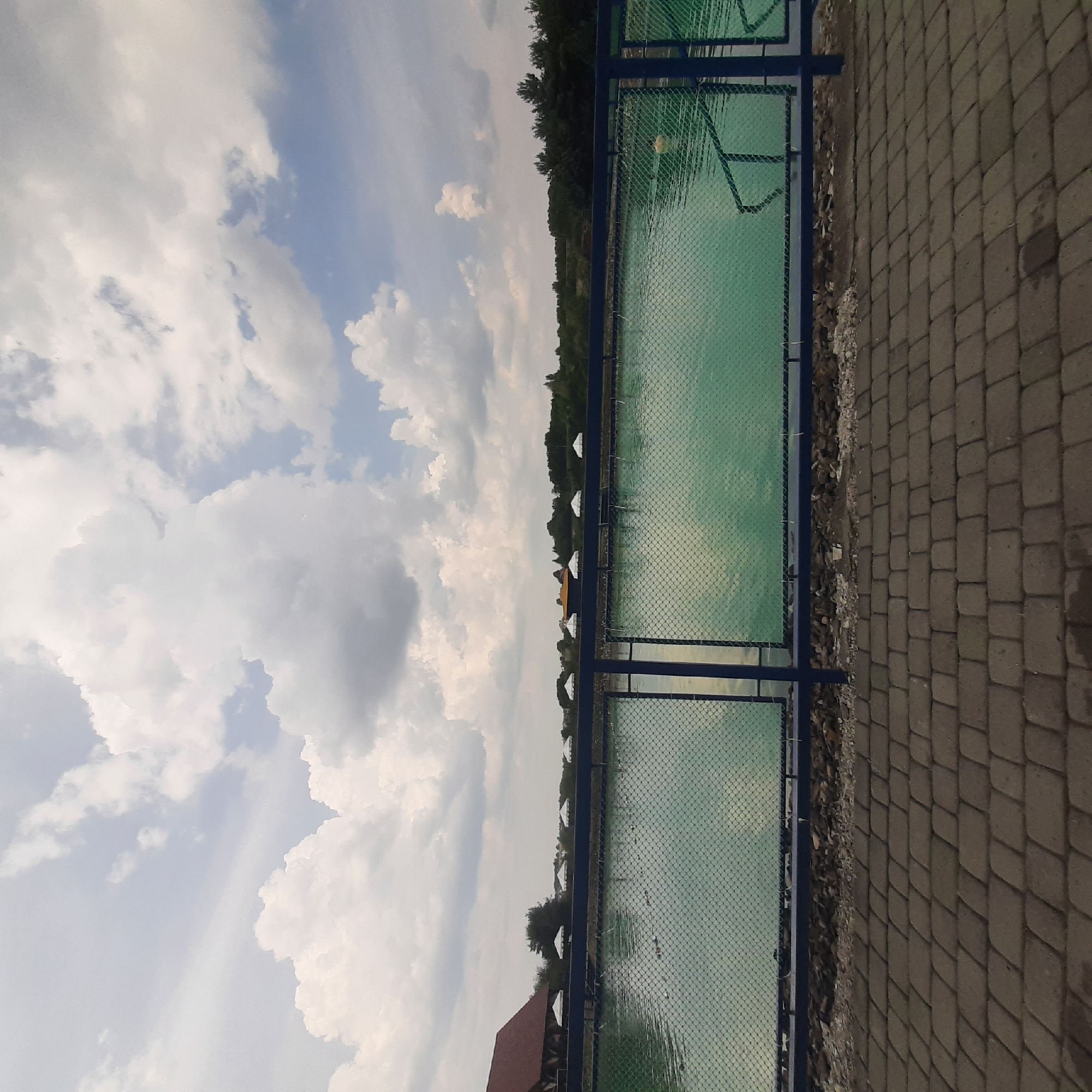 The image depicts a man-made pond in Dubrivka, Ukraine, likely an aquaculture or fishing facility, based on the presence of what appear to be fish cages or netting in the water. A dark blue metal fence with green mesh separates the viewer from the pond. The pond's water is a vibrant turquoise color, reflecting the cloudy sky above. Along the far bank of the pond, several white canopy tents are visible, suggesting an area for visitors or vendors. Behind the tents, a dense line of trees forms the horizon. To the left, a rustic building with a red roof is partially visible. The foreground consists of a paved walkway made of interlocking gray bricks. The overall scene is naturalistic, with the pond, vegetation, and sky dominating the landscape. There are no people visible in the image.