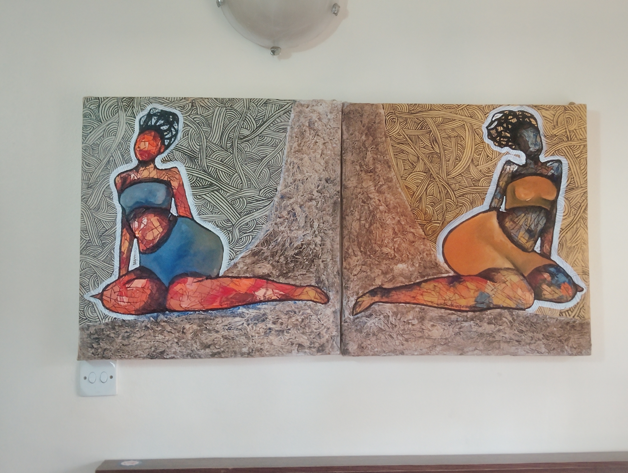 The image shows two abstract paintings of women displayed on a white wall. The paintings are hung side-by-side, with the left painting featuring a woman in a blue garment against a black and white patterned background. The right painting depicts a woman in an orange top and orange lower body against a gold patterned background. Both paintings have a textured, rough surface in the lower half, resembling bark or dried mud. Below the left painting, a white electrical outlet with two round buttons is visible. The ceiling light fixture is partially visible at the top of the image. The overall impression is that of an indoor art display. This scene is located in Alagbado, Nigeria.