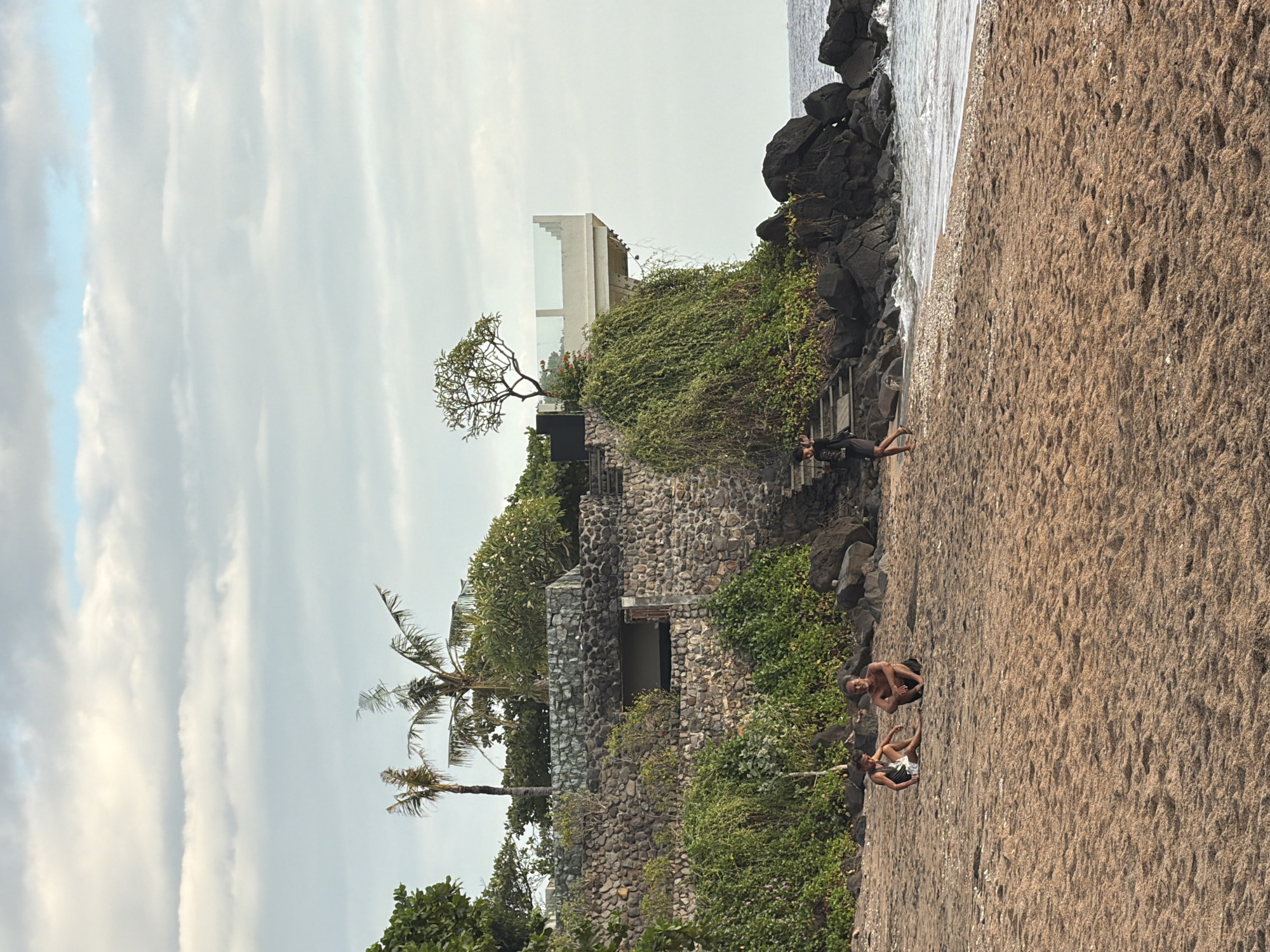 The image depicts a beach scene in Abang, Indonesia, with a stone building on a cliff overlooking the ocean. In the foreground, on the sandy beach, two individuals are seated, and another person is walking near the water. The building on the cliff is partially covered in lush green vegetation, and a modern observation deck with a glass railing extends from the cliff face. Palm trees are visible behind the building. The ocean is calm, with gentle waves lapping at the shore. The sky is partly cloudy, with soft light suggesting either early morning or late afternoon.