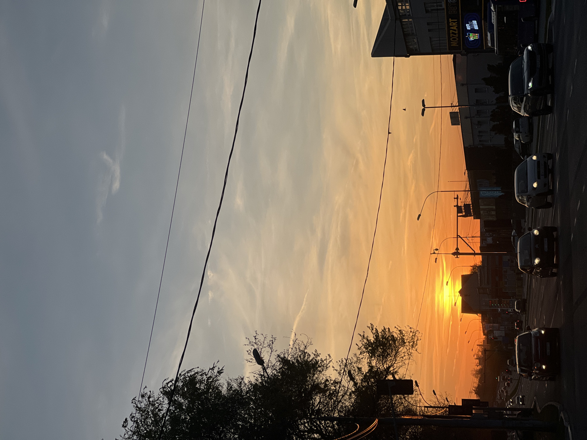 Captured at Palilula Urban Municipality, Serbia, the image presents a street view at sunset. Several cars are on the road, their headlights illuminating the dusk. Streetlights are interspersed along the road. The sky transitions from pale blue to a fiery orange. Trees frame the lower portion of the image, with utility wires cutting across the sky. A building with a “MOZZART” sign is visible along the street.