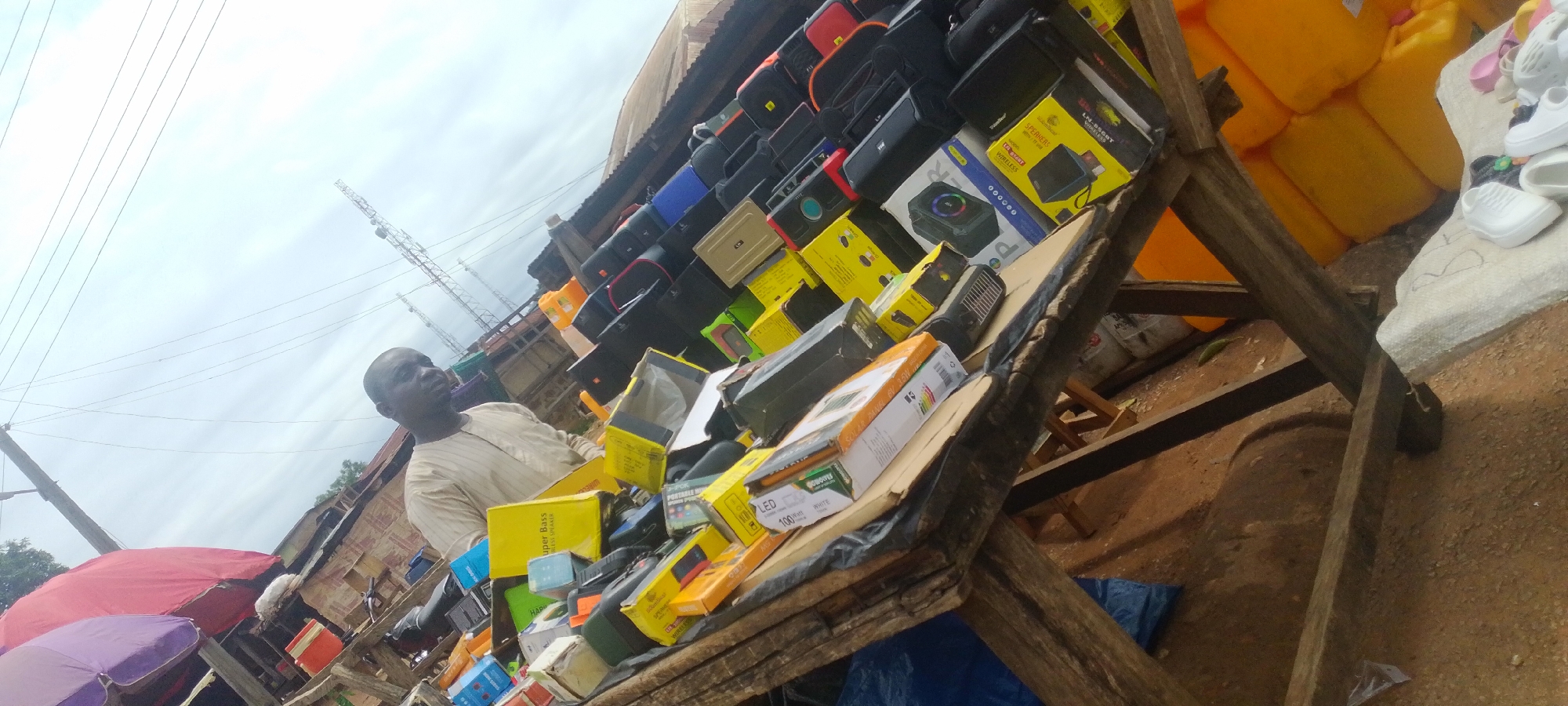 The image shows a man standing behind a stall laden with various electronic devices and accessories, likely in a marketplace in Baissa, Nigeria. The stall features a wide assortment of items, including portable speakers, headphones, chargers, and other electronic gadgets, many of which are in their original packaging. The packaging is predominantly yellow and black, with some items also in white.
The man, dressed in a light-colored traditional outfit, appears to be a vendor attending to his stall. He is looking slightly upwards and to the right, his expression neutral. The stall itself is constructed from rough wooden planks, suggesting a rustic market setting. Behind the stall, large yellow plastic containers are stacked, and further back, a building with a corrugated iron roof is visible. Power lines and two tall communication towers are prominent in the background, indicating an urban or semi-urban environment.
The sky is overcast, suggesting a cloudy day, possibly late morning or afternoon given the lighting. The ground is a mix of dirt and sand. To the left of the stall, a red and a purple umbrella are partially visible, adding to the market atmosphere. A few pairs of white shoes are also visible on a surface to the right.
Visible text on the packaging includes "Super Bass", "PORTABLE SPEAKER", "LED 100W WHITE", and "LN-BSGT BLUETOOTH". The overall impression is a busy, open-air marketplace where electronics are being sold.