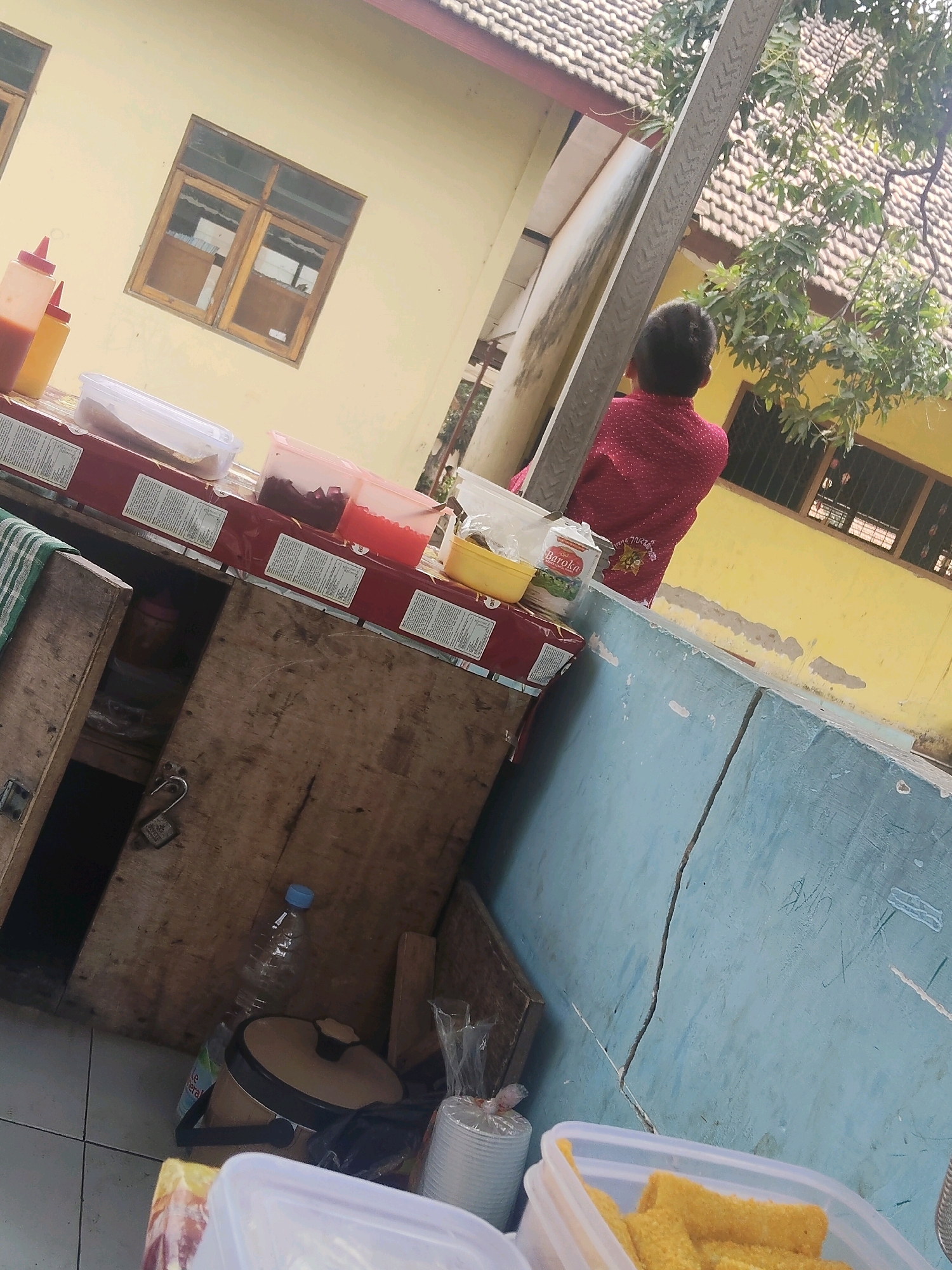 The image depicts an outdoor scene, likely in Payungrejo, Indonesia, featuring a makeshift food stall. In the foreground, a wooden cabinet, weathered and secured with a padlock, holds various food items and containers. To the left, a green and white checkered cloth is draped over the cabinet. On top of the cabinet, several clear plastic containers hold food ingredients, including what appears to be red jelly or candy, and a white powdered substance. A yellow plastic container and two squeeze bottles, one containing a red liquid and the other a yellow liquid, are also visible.
In the background, a yellow building with a tiled roof stands. A window with wooden frames is visible on the building. To the right, a young person, seen from behind, is wearing a red patterned shirt. They are standing near a concrete structure painted light blue. Lush green trees with foliage partially obscure the building and the sky. A plastic bottle and a small trash can are placed on the ground near the cabinet. In the lower right corner, a clear plastic container is filled with rectangular, breaded food items, possibly snacks. The overall scene suggests a casual, outdoor food vending operation.