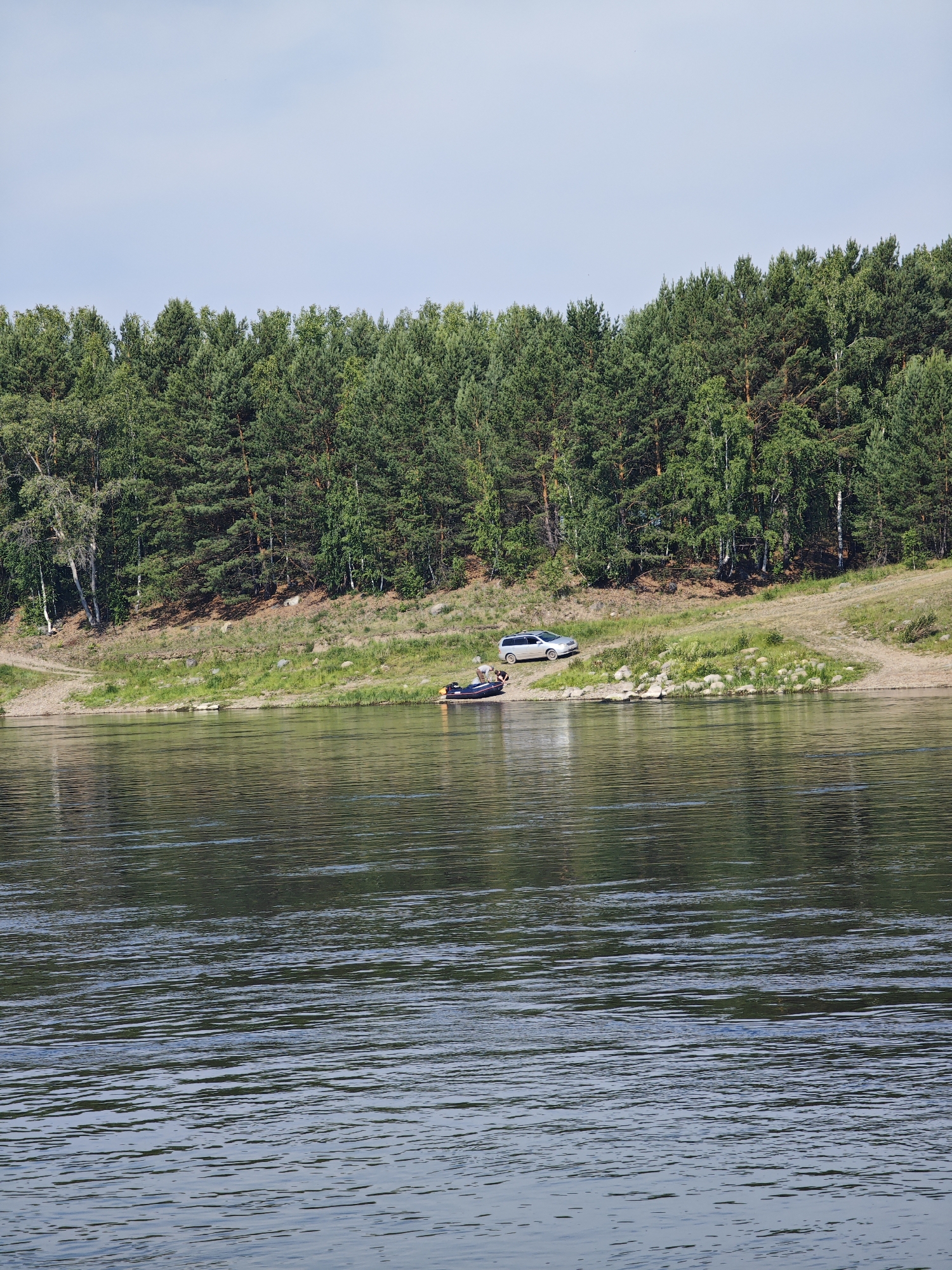 A wide, calm river flows in the foreground, reflecting the trees and sky. The riverbank on the far side is lined with a dense forest of pine and birch trees. Two silver-colored cars are parked on the grassy bank near the edge of the forest. A small inflatable boat with two people is visible on the water, close to the shore. The sky is clear and pale blue. The scene depicts a tranquil natural setting on a sunny day.