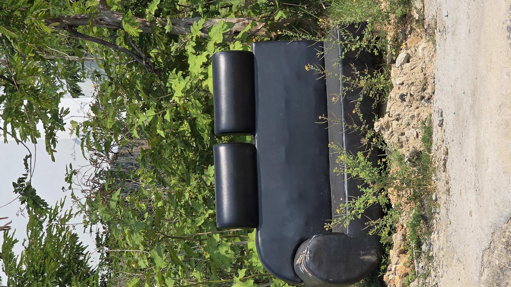 A black, modern-style sofa sits outdoors in Bắc Nha Trang, Vietnam. The sofa, featuring two cylindrical back cushions and a rounded end, is positioned on a patch of dirt and gravel alongside a concrete surface. Green vegetation surrounds the sofa, with trees and bushes visible in the background. The lighting is bright, suggesting a sunny day. No people are visible in the scene.