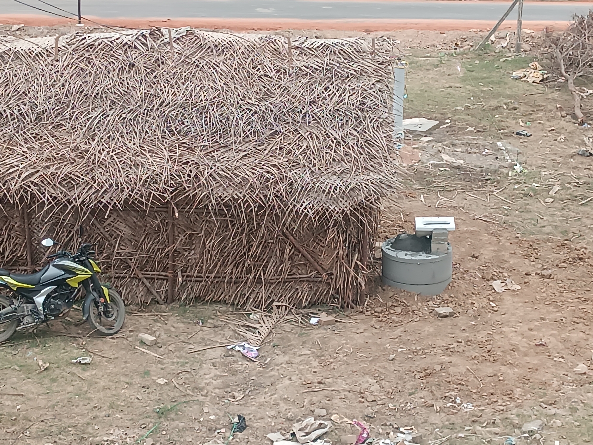 A motorcycle is parked next to a hut made of dried palm fronds in Senneerkuppam, India. A concrete well with a metal lid is located near the hut. The ground is dirt and littered with debris. A road and foliage are visible in the background.