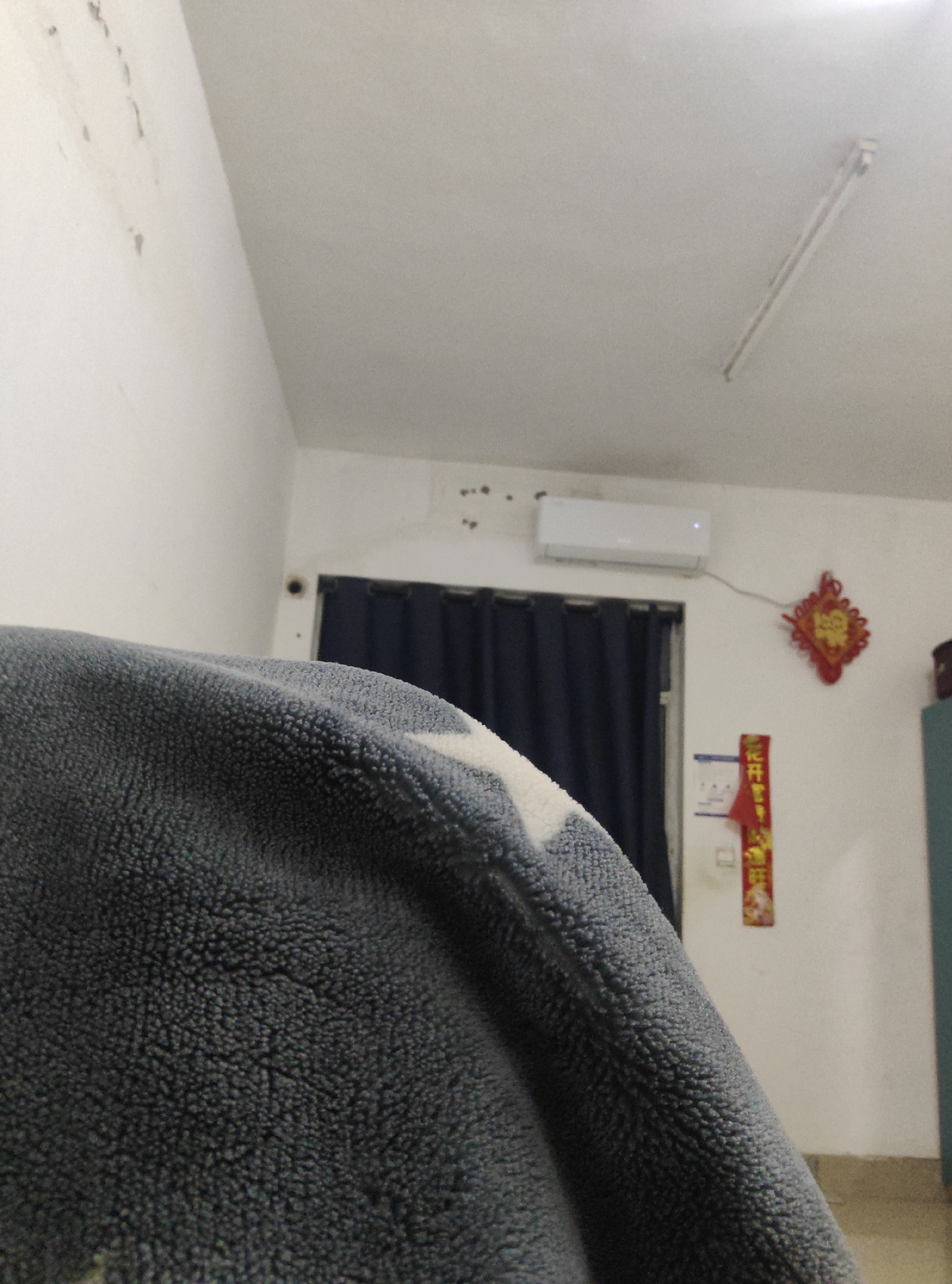 A close-up, low-angle, indoor shot depicts a fuzzy, dark grey blanket in the foreground. A white, star-like pattern is visible on the blanket. In the background, a white wall displays some water stains and possibly soot marks. A dark blue curtain hangs behind a white air conditioner unit mounted on the wall. To the right of the air conditioner, a red, diamond-shaped Chinese ornament is attached to the wall, with Chinese characters in red and gold. Below this ornament, a vertical red banner with gold Chinese characters is also affixed to the wall. A fluorescent light fixture is visible on the ceiling. The scene appears to be a domestic interior, possibly a bedroom, with no specific indication of the Chongqing High-tech Zone, China, other than the prompt's request.