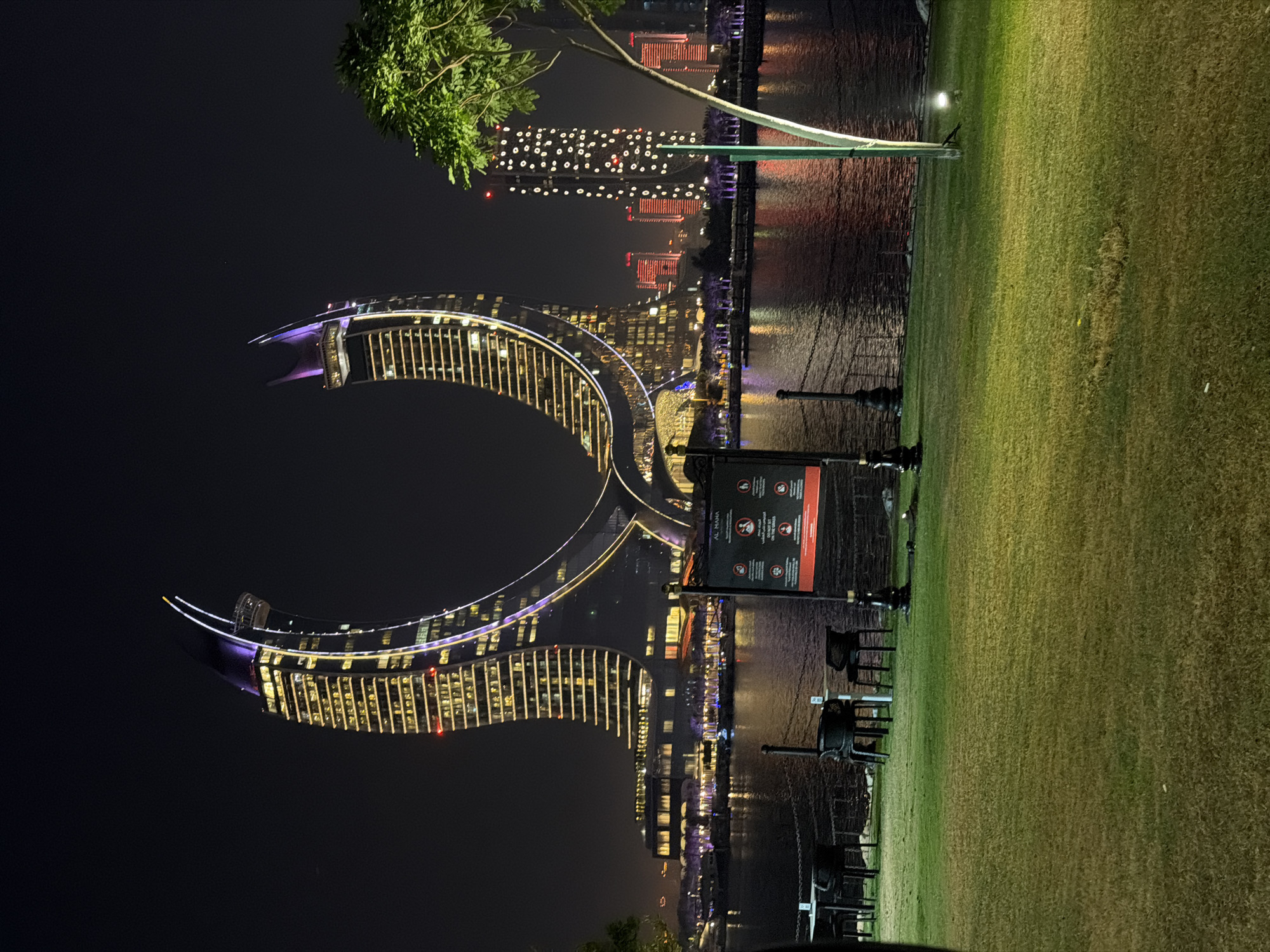 The image depicts a nighttime scene at a waterfront location, likely a park or a public space, in an urban environment. In the background, two distinct, futuristic-looking buildings dominate the skyline. The larger of the two buildings has a distinctive curved, horseshoe-like shape and is illuminated with bright yellow lights outlining its structure, with purple lights accentuating its upper curves. To the right of this building, a cluster of taller, more rectangular buildings are visible, also lit up with various patterns of white and red lights. The buildings are reflected in the dark water of a canal or bay that separates the foreground from the background.
In the foreground, a well-maintained lawn is visible, illuminated by soft, green lighting. Several outdoor tables and chairs are arranged on the lawn, suggesting a seating area for a cafe or restaurant. A dark-colored information board with text and icons is positioned on a pole near the tables. To the right of the lawn, a tree with a brightly lit trunk is visible, and further to the right, a section of the waterfront with a light post is seen. The overall impression is one of a modern and vibrant cityscape at night, with a focus on architectural illumination and recreational outdoor spaces.
