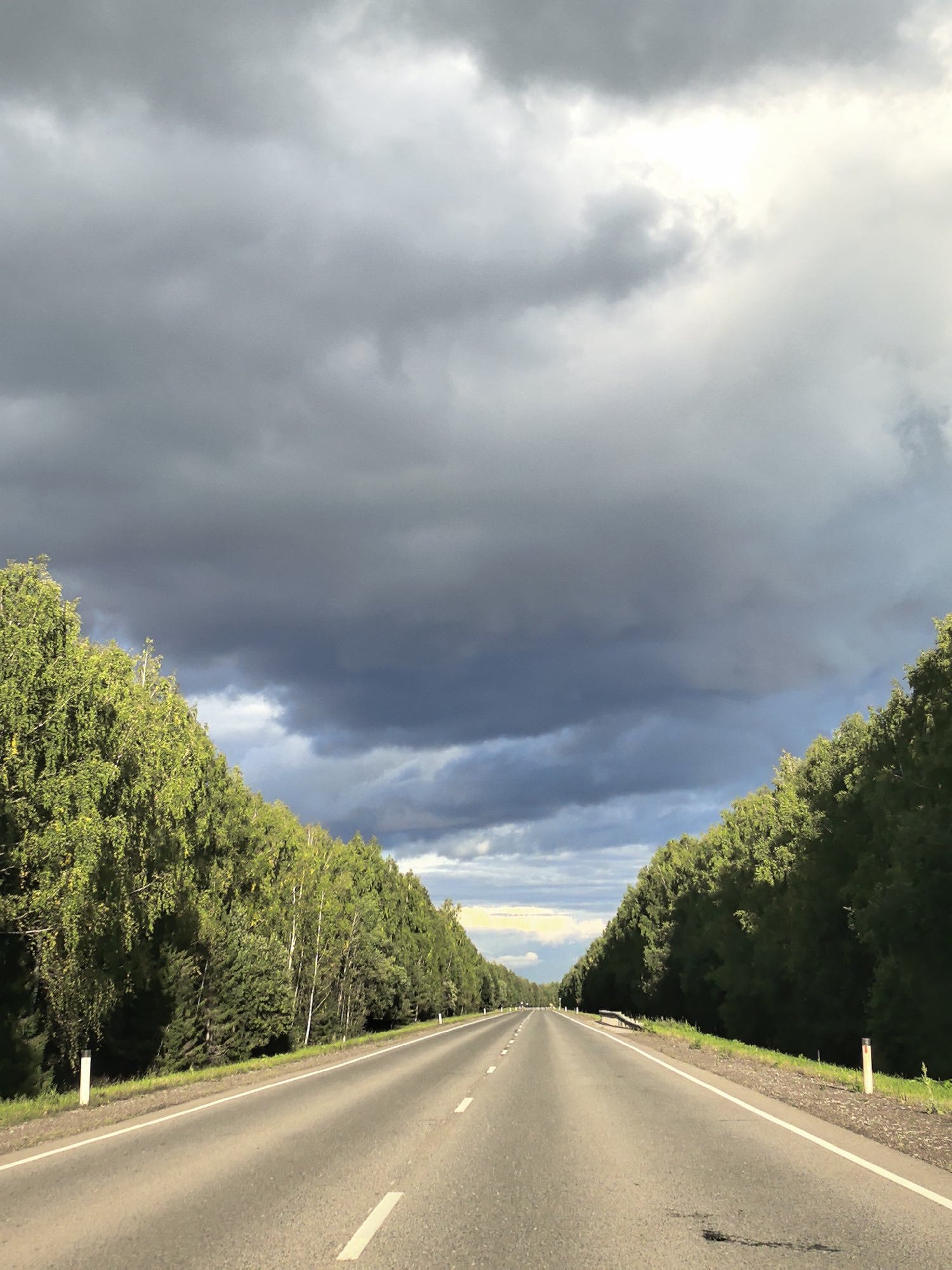 An asphalt road stretches into the distance, flanked by dense green trees on either side. The sky above is dramatic, filled with heavy, dark storm clouds that partially obscure a bright, light-filled area in the distance. The road is marked with white lines, indicating lanes. There are no people or animals visible in the image. The scene appears to be a rural or semi-rural highway in Shabury, Russia, with the impending storm creating a moody atmosphere.