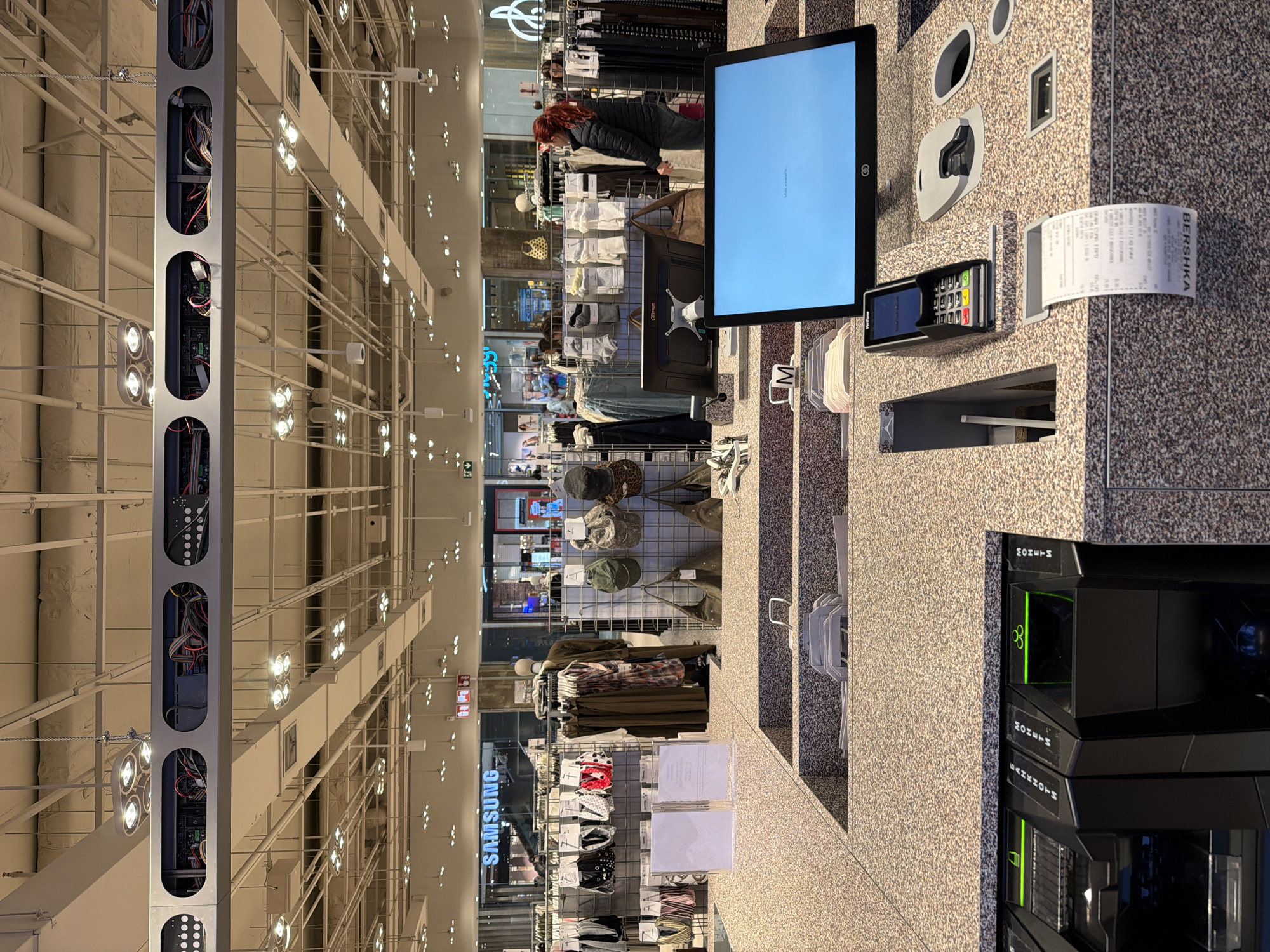 The image depicts an interior scene, likely a retail store located in Sofia, Bulgaria, based on the “BERSHKA” signage on a receipt. The store appears to be a clothing or accessories shop, as evidenced by displays of items like hats, socks, and clothing racks in the background. A woman with red hair is visible browsing merchandise. A self-checkout station dominates the right side of the image, featuring a digital display screen, a card reader, a receipt, and automated return machines. The lighting is bright, with rows of lights illuminating the open ceiling structure. A "SAMSUNG" sign is visible in the background, suggesting the presence of an electronics department or promotional display.
