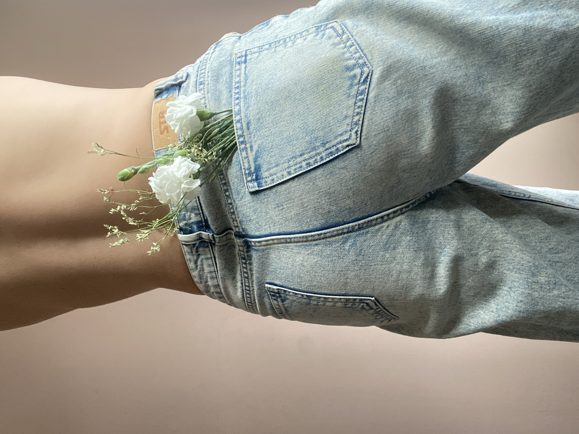 A photograph features the lower back and rear of a person wearing acid-wash blue jeans, with a small bouquet of white flowers placed in one of the back pockets. The person is positioned against a light pink or beige background. No specific details indicate the location being Chaikovsky, Russia.