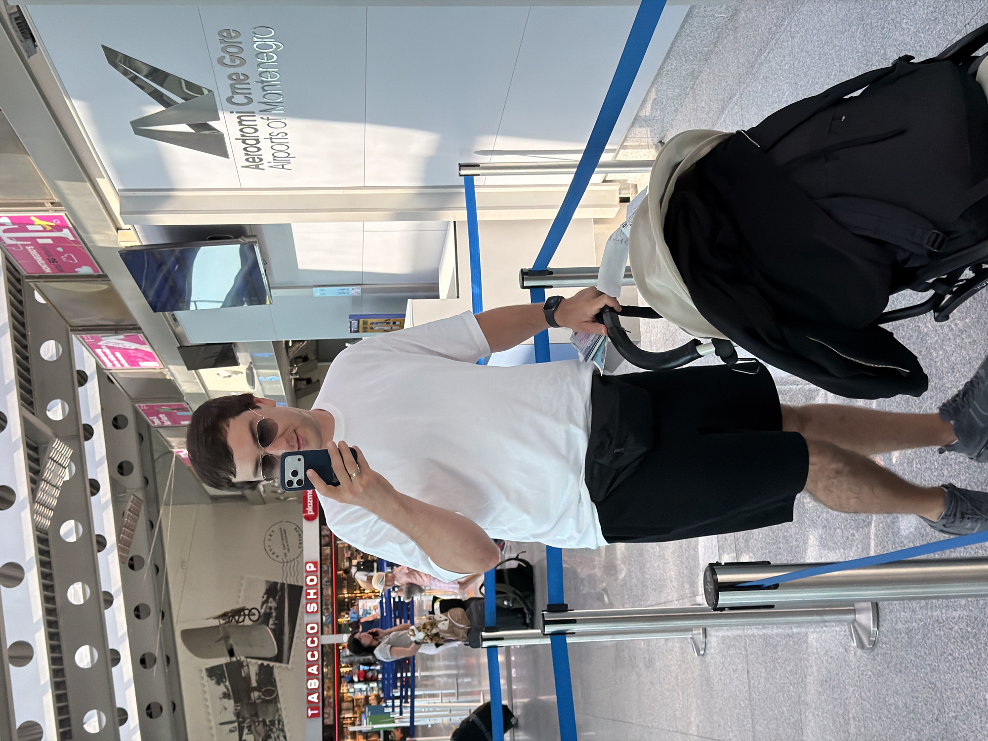 A man stands in an airport terminal, holding a smartphone and a stroller. He is wearing a white t-shirt, black shorts, and a black fanny pack. He is also wearing sunglasses and a watch. The stroller is black and has a beige canopy. In the background, there are shops, signs, and other travelers. The "Aerodromi Crne Gore, Airports of Montenegro" sign indicates the location is likely Podgorica Airport in Donja Cijevna, Montenegro. The man is positioned near a queue barrier.