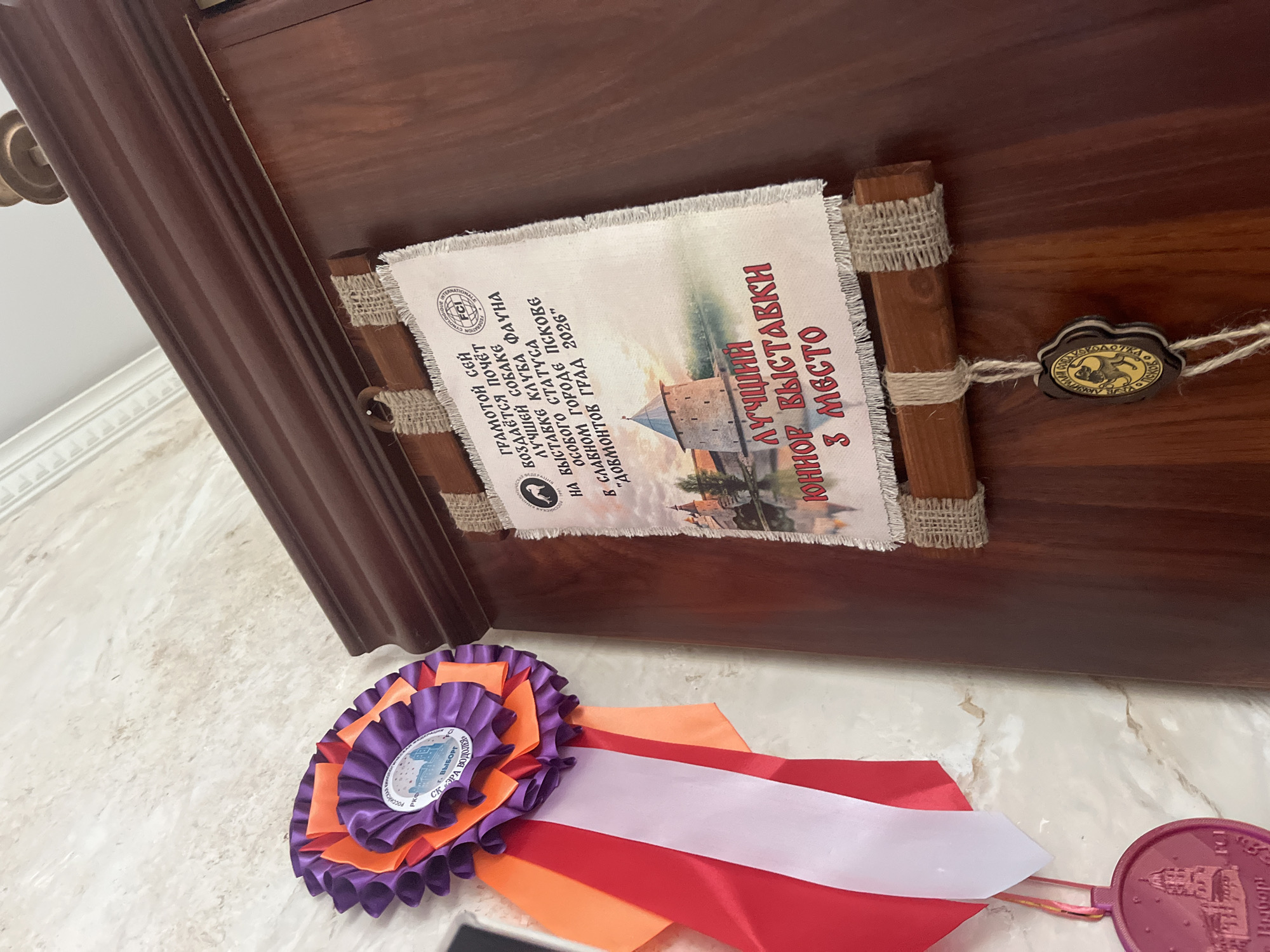 The image displays a close-up view of awards displayed on a wooden surface. On the left, a ribbon rosette in purple and orange with red and white streamers hangs against a marble-patterned wall. Below this, a dark purple medal with an engraved building is visible. To the right, a diploma or certificate is attached to the wooden surface with twine and burlap strips. The diploma features an illustration of a tower and text, including "FCI" and Cyrillic characters. A circular wooden seal with an image of a cat and Cyrillic text is suspended below the diploma. The wooden surface appears to be part of a cabinet or piece of furniture with a dark, polished finish. The overall scene suggests an indoor display of dog show or competition awards, possibly in a home or a specialized venue. The location in Vladimirovka, Russia is not explicitly indicated by any visible elements within the provided image.