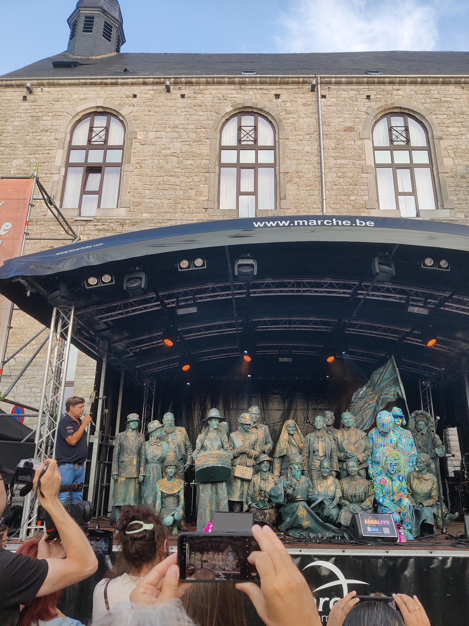 The image depicts an outdoor event in the city of Marche-en-Famenne, Belgium, likely taking place in a public square or plaza in front of a stone building with arched windows. In the foreground, several individuals are holding up cameras and smartphones to capture the scene. A man in a dark blue polo shirt and jeans is speaking into a microphone on a stage.
The main focus of the image is a group of costumed performers on a stage, appearing to be reenacting historical or artistic figures. These performers are adorned in metallic, bronze-like costumes, with one individual dressed in a brightly colored, mosaic-patterned outfit, possibly representing a modern interpretation or contrast. The figures are posed in various stances, some holding props like a flag and a bucket labeled "ICE CREAM." A sign that reads "ABBALIVE" is visible on the stage.
The backdrop features the stone facade of a building with a dark slate roof and a bell tower. The overall atmosphere suggests a cultural performance or festival. The website "www.marche.be" is visible on the awning of the stage, confirming the location in Marche-en-Famenne. The sky above is partly cloudy.