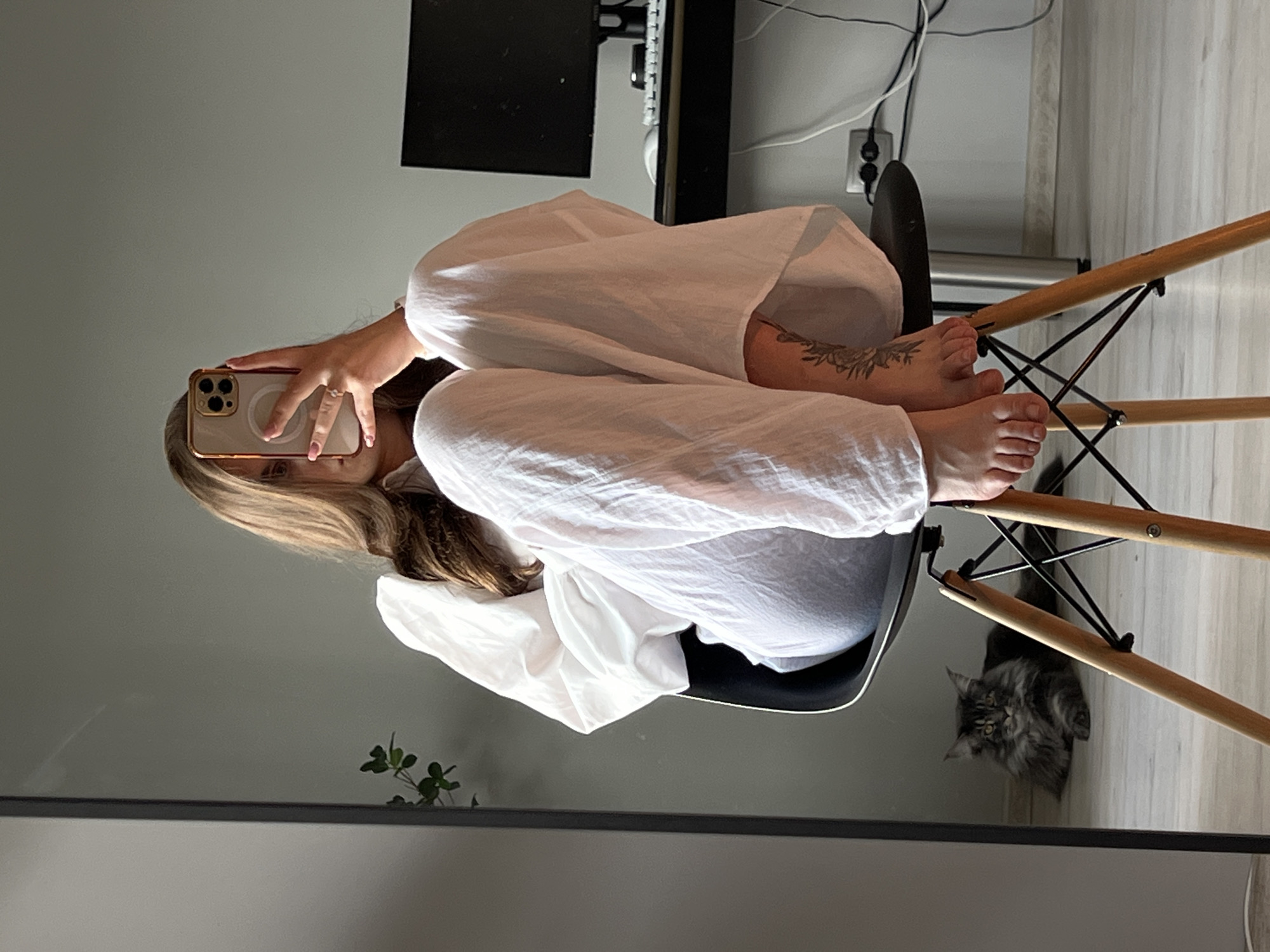 An indoor scene captures a woman taking a mirror selfie, with a cat resting nearby. The woman, with long light brown hair, sits on a black chair with splayed wooden legs. She wears a white, loose-fitting long-sleeved shirt and matching loose white trousers. Her face is obscured by a light-colored smartphone, featuring a gold-toned case and triple camera, which she holds with her left hand, revealing light pink nail polish and a ring on her ring finger. Her bare feet are visible, one resting on the chair, the other dangling, exposing a dark, possibly floral or leafy, tattoo on her lower right leg.
To the left, a large rectangular mirror with a thin black frame reflects a dark grey Maine Coon cat lying on a light wooden floor, looking towards the viewer. A small green potted plant is also partially visible in the mirror's reflection. To the right of the woman, a dark desk holds a black computer monitor, a white keyboard, and a white mouse. Multiple cables, both white and black, are visible near a power outlet strip on the light grey wall behind the desk. The room has light grey walls and a light wooden floor. The specific city location of Ternopil, Ukraine, cannot be determined from the visual information in the image.