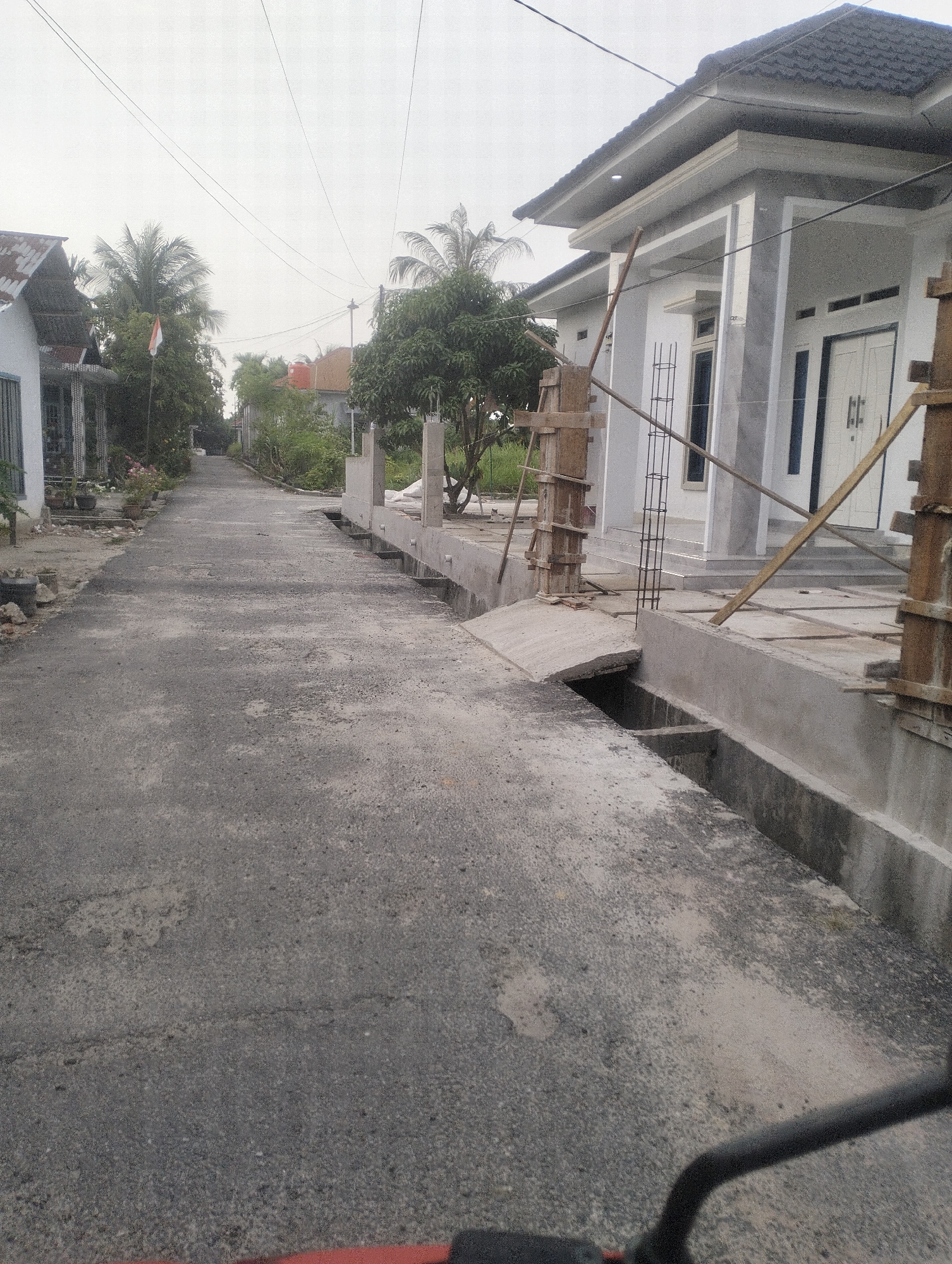 The image depicts a paved road in Tambusai Batang Dui, Indonesia, with a new residential building under construction on the right side of the frame. The road is asphalt and appears to be in a residential area, as evidenced by the houses on the left side of the road and the presence of palm trees and other vegetation. The building under construction is a modern, multi-story structure with a gray exterior and a dark tiled roof. The construction site is active, with scaffolding and building materials visible. The weather appears overcast. The image is taken from a low angle, possibly from a vehicle.