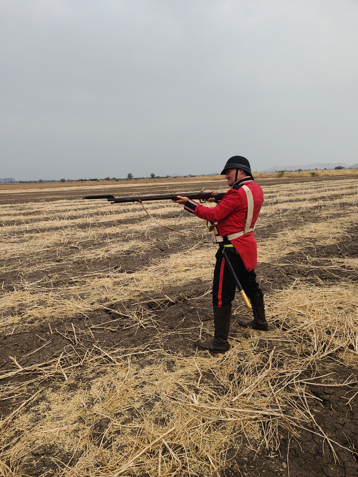 A male figure stands in a dry agricultural field in Kottalapalli, India, under an overcast sky. The ground consists of dark brown, cracked soil sparsely covered with rows of light brown, dry crop stubble. In the distant background, a flat landscape transitions to low-lying hills or mountains beneath the grey horizon. The man, appearing Caucasian, wears a historical military uniform: a black pith helmet, a red long-sleeved jacket with white shoulder straps and dark cuffs, dark trousers with red side stripes, and dark knee-high boots. A light-colored belt encircles his waist, from which a yellow/gold-colored object and a fluorescent yellow-green band are suspended on his left hip. He holds a long, dark musket or rifle, fitted with a bayonet, in a horizontal aiming position, his hands gripping the weapon. The rifle also features a light-colored sling. He is facing slightly towards the left side of the frame.