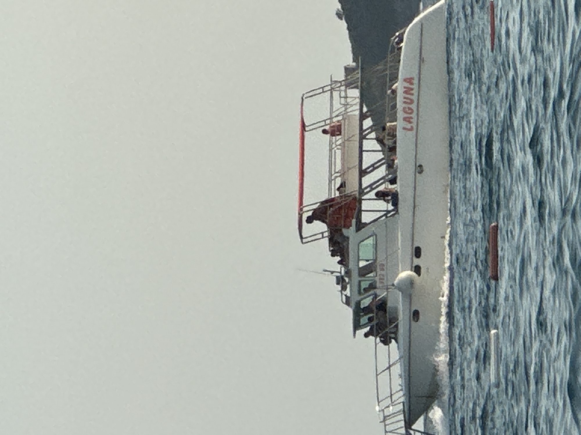 A white motor yacht named "LAGUNA" is cruising on choppy blue water. The yacht has multiple levels with people visible on both the lower and upper decks. The boat is white with a red trim and a red canopy over the upper deck. In the foreground, there are two cylindrical buoys floating on the water, one white and one orange. In the background, a green, tree-covered hill slopes down towards the water, with a small tree visible at its peak. The sky is a pale, hazy blue. The image is taken from a low angle, looking up at the yacht. The location is the waters near Boreti, Montenegro.