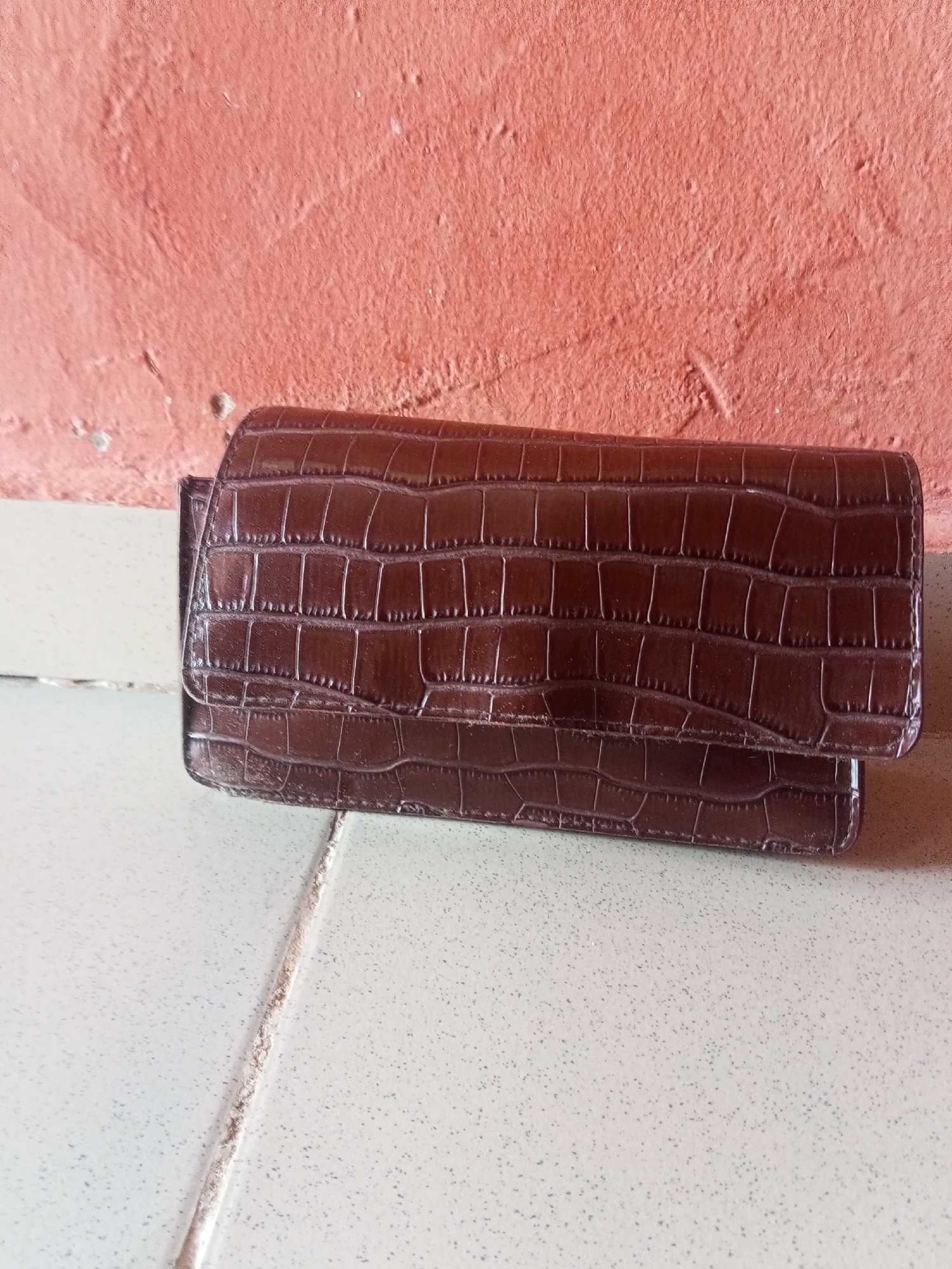A brown faux alligator skin wallet is laid on a white tiled floor. The wallet is long and flat, with a folded over flap secured by a hidden clasp. The wallet is positioned diagonally across the frame, with the top edge closer to the viewer. Behind the wallet is a textured, salmon-colored wall with visible imperfections. The lighting appears to be natural, casting soft shadows on the tiled surface. There are no people or other objects present in the image.