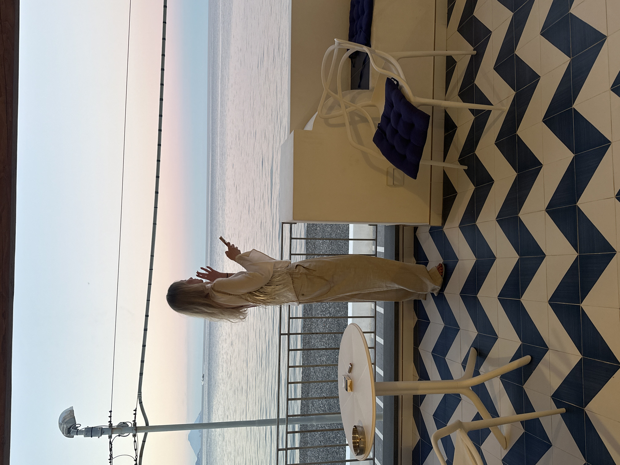 A woman with long blond hair, dressed in beige clothing, stands on a balcony overlooking the sea in Lipari, Italy, holding a smartphone. She is taking a picture or video. The balcony is adorned with blue and white chevron-patterned tiles. A white round table with a cigarette ash tray is present, along with white chairs. In the background, a serene seascape stretches to the horizon under a sky with hues of pink and blue. An electrical pole is visible on the left side of the frame.