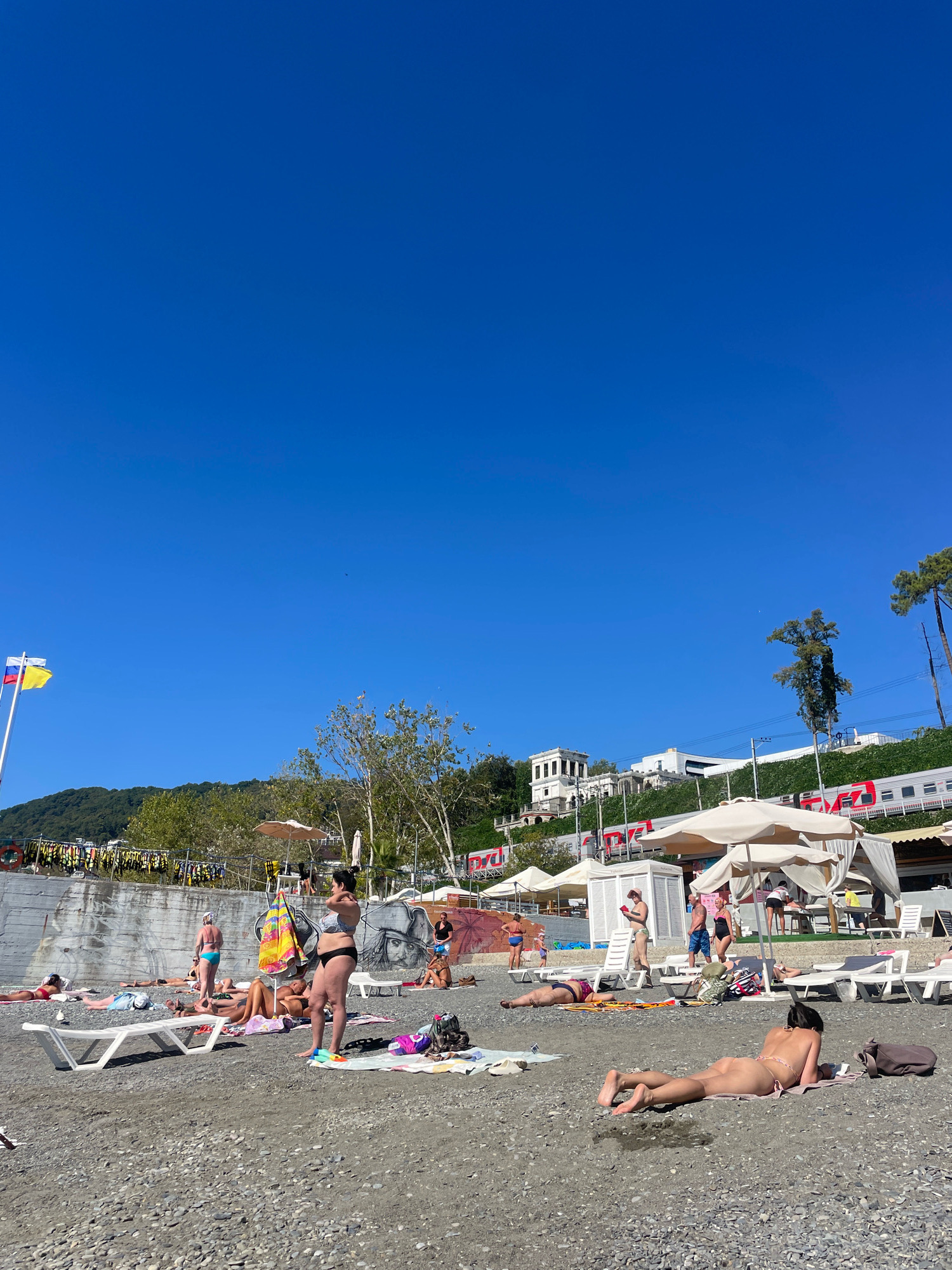 On a sunny day, a group of people are sunbathing on a pebble beach in Sochi, Russia. The beach is situated near a cliff with a wall covered in graffiti. A train is visible on tracks above the beach, and several white tents with umbrellas provide shade. The sea is not visible in the frame. In the foreground, several individuals are lying on towels and loungers, some wearing swimwear and others nude. A woman in a bikini stands near a colorful umbrella, while another woman is lying on her stomach, facing away from the camera. Flags, including the Russian and Ukrainian flags, are flying on poles in the background. The sky is clear and blue.