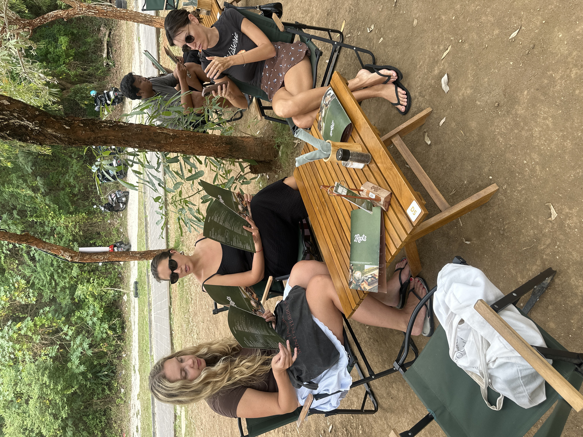 Three women are seated at a wooden table outdoors, with trees and foliage in the background. The woman on the left, with blonde hair, wears a brown t-shirt and a blue and white striped skirt, and is looking down at a menu. A dark tote bag with "Saint-Petersburg" printed on it is on her lap. The woman in the middle wears a black dress and black sunglasses, and is also looking at a menu. The woman on the right wears a black t-shirt with text, a brown patterned skirt, and sunglasses. She is holding a smartphone and looking at it. A wooden table in front of them holds menus, salt and pepper shakers, and cups with napkins. In the background, several motorcycles are parked, and a man is seated behind the table, also looking at a menu. The ground is dirt, with some scattered leaves. This scene appears to be at a casual outdoor dining establishment in Kuta Selatan, Indonesia.