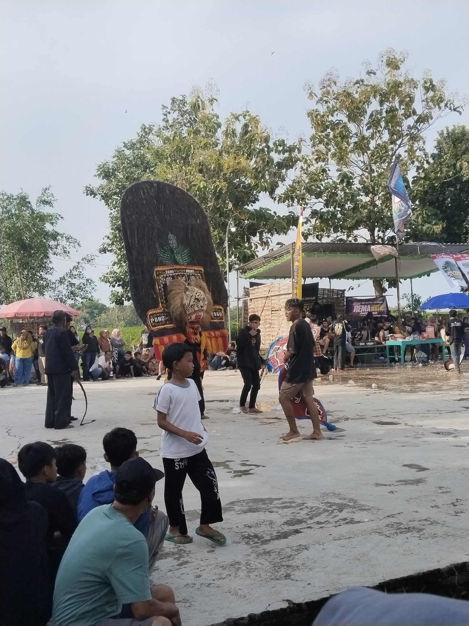 The image shows a gathering of people in Mojodoyong, Indonesia. At the center is a large, elaborate headdress, likely part of a traditional performance. Two people are standing beside it. One person stands to the left, carrying a whip, while another performs in front of the headdress. People are seated on the ground in the foreground, watching the performance. In the background, more spectators are gathered under a tent-like structure. The ground is paved with cracked concrete, and trees are visible in the background. A bird is visible flying in the distance. Banners and flags are visible next to the tents.