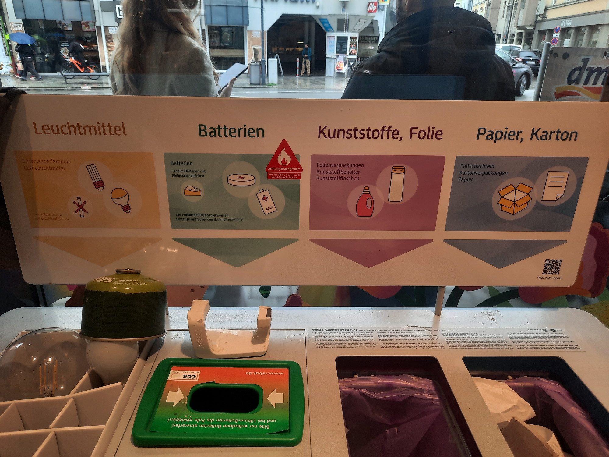 A close-up, slightly angled indoor shot captures a recycling information sign and collection bins. The sign, mounted above the bins, is divided into four sections: "Leuchtmittel" (Lighting), "Batterien" (Batteries), "Kunststoffe, Folie" (Plastics, Foil), and "Papier, Karton" (Paper, Cardboard). Each section illustrates acceptable items with simple graphics. The "Batterien" section includes a warning symbol for fire hazard. Below the sign, various recycling bins are visible. On the left, a white plastic organizer holds a clear glass bulb and a green camping gas canister. In the center, a white plastic holder with a curved handle is positioned next to a green collection bin with an orange label featuring the CCR logo and instructions in German about battery disposal. To the right, a larger black bin is partially visible, containing purple trash bags and some white paper waste. The background shows a street scene through a glass window, with buildings, vehicles, and pedestrians. The presence of a "dm" drugstore signage in the background suggests the location is within Rosenheim, Germany, likely at a public collection point or a store offering recycling services.