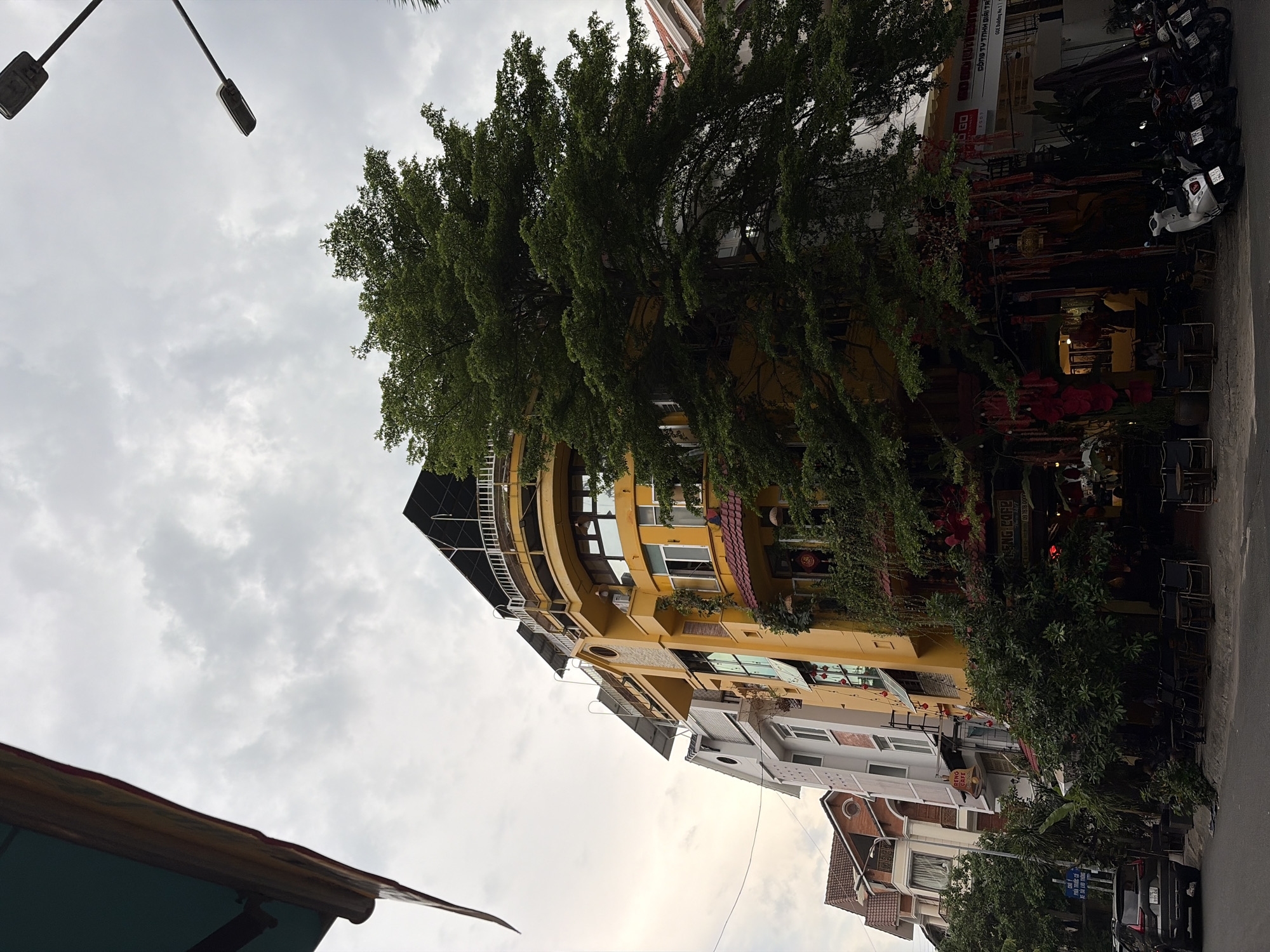 This is a street-level, eye-level shot taken in Thủ Đức, Vietnam, under a cloudy sky. The scene is dominated by a vibrant yellow building with rounded corners, decorated with lush greenery cascading down its facade. The building has multiple stories, featuring balconies with white railings and numerous windows. To the left of the yellow building, a row of narrower, lighter-colored buildings with traditional sloped roofs are visible. In front of the buildings, a sidewalk area is set up with tables and chairs, suggesting an outdoor cafe or restaurant. Several parked motorcycles are lined up on the street. The street itself is paved, and a dark-colored SUV is parked on the left side. A tall, leafy green tree extends from the right side of the frame, partially obscuring the yellow building and reaching towards the sky. Overhead, two streetlights are visible against the overcast sky. The overall impression is that of a bustling urban environment with a strong presence of nature integrated into the architecture.