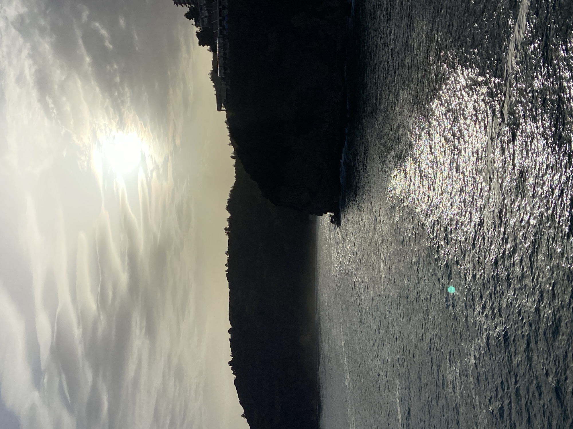 The image depicts a coastline with a dark, steep hillside meeting the sea. The sky is overcast with a bright sun partially obscured by clouds. In the foreground, the water surface is reflective and shimmering. Buildings can be seen silhouetted at the top of the cliff. Based on the landscape, this image is likely in the city of Budva, Montenegro.