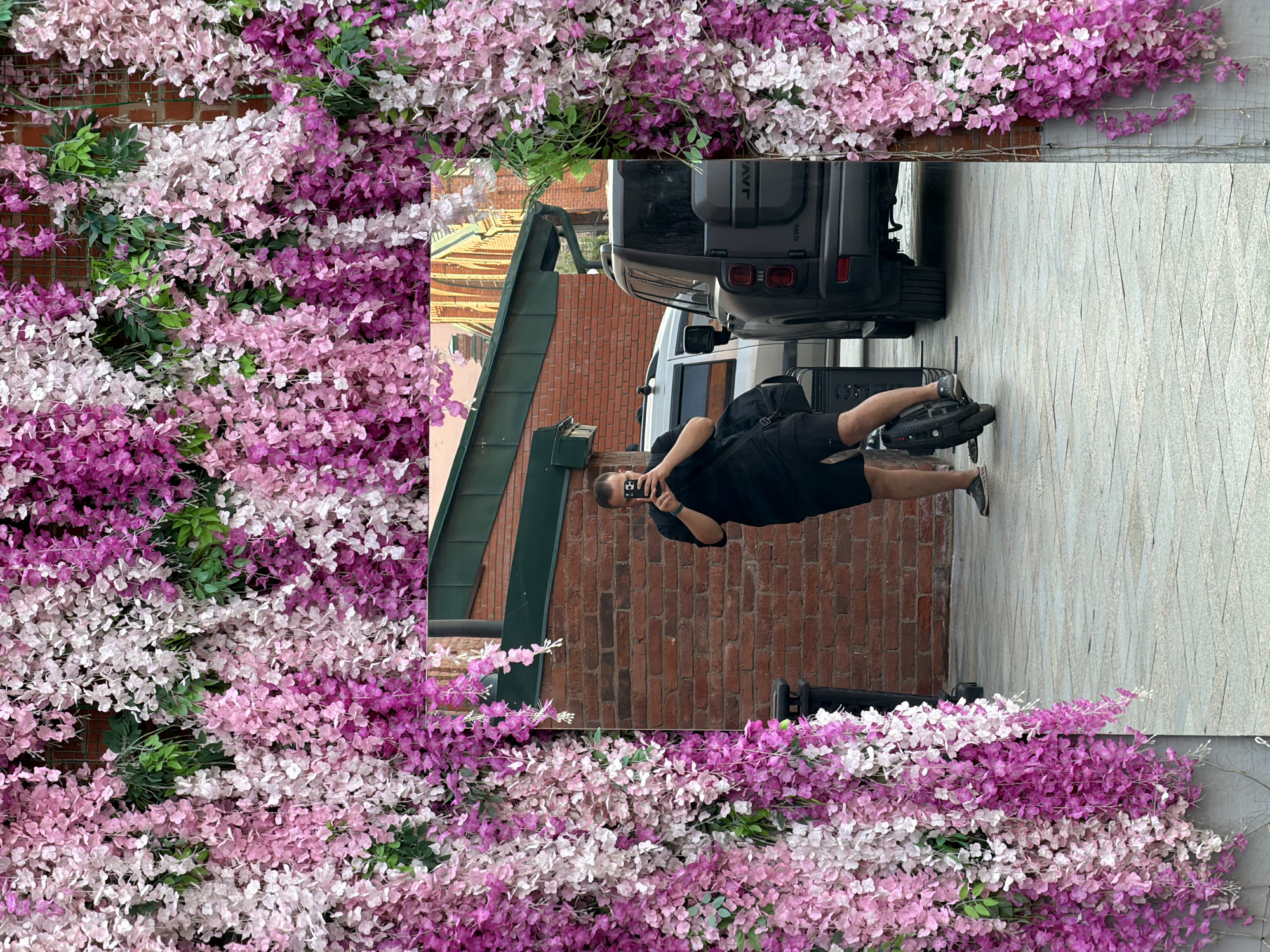 A man in black clothing is positioned to the right of the frame, holding a smartphone and seemingly taking a picture. He is riding what appears to be a single-wheel electric skateboard. Behind him is a parked vehicle, partially visible. The scene is framed by floral decorations, predominantly in shades of pink and purple. A brick wall and building elements can be seen in the background. The location may be Moscow, Russia, but there is no explicit confirmation.