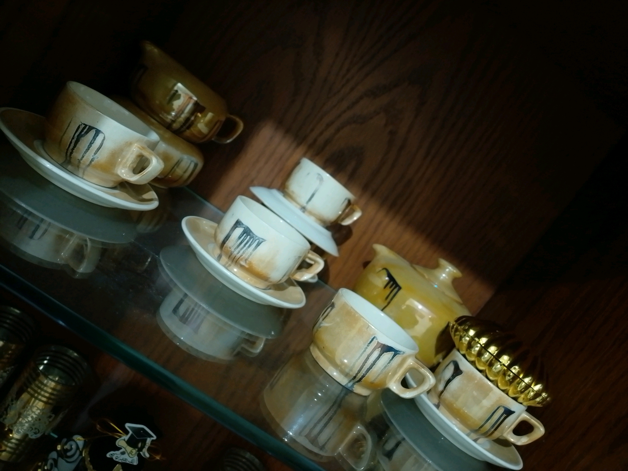The image shows a collection of tea cups and saucers, along with a teapot, displayed on a glass shelf in what appears to be a cabinet with a wooden background. The tea set features a light beige color with dark, abstract vertical streaks. Some cups are stacked, and others are paired with saucers. A distinctive golden, ribbed lid is placed on top of one of the cups. In the lower left corner, there are also some decorative items, including what looks like a graduation cap charm. The lighting suggests an indoor setting, possibly a home or a shop, and there are no people or discernible city landmarks visible. The scene does not provide any specific geographical indicators that would place it in Amman, Jordan, or any other city.