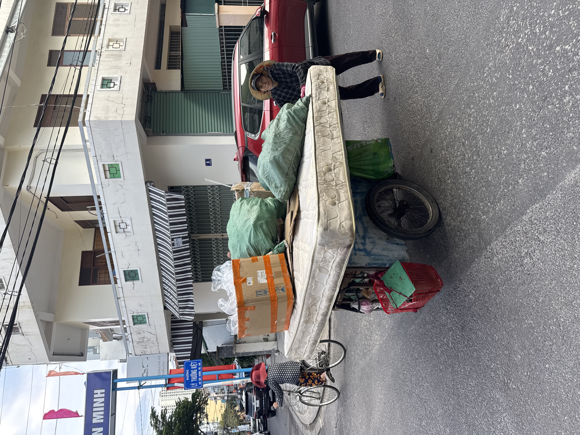 In Nha Trang Ward, Vietnam, on a paved street, a woman is pushing a cart loaded with a mattress and several large green bags of what appears to be trash or recyclables. The woman is wearing a plaid shirt, dark pants, a straw hat, and sandals. She has her right hand on the cart. To the left of the cart, a person on a bicycle wearing a red hat and patterned clothing is riding away. In the background, a red SUV is parked, and a multi-story building with a balcony and green metal shutters is visible. A street sign for "ĐƯỜNG LÝ THƯỜNG KIỆT" is also present. The sky is overcast.