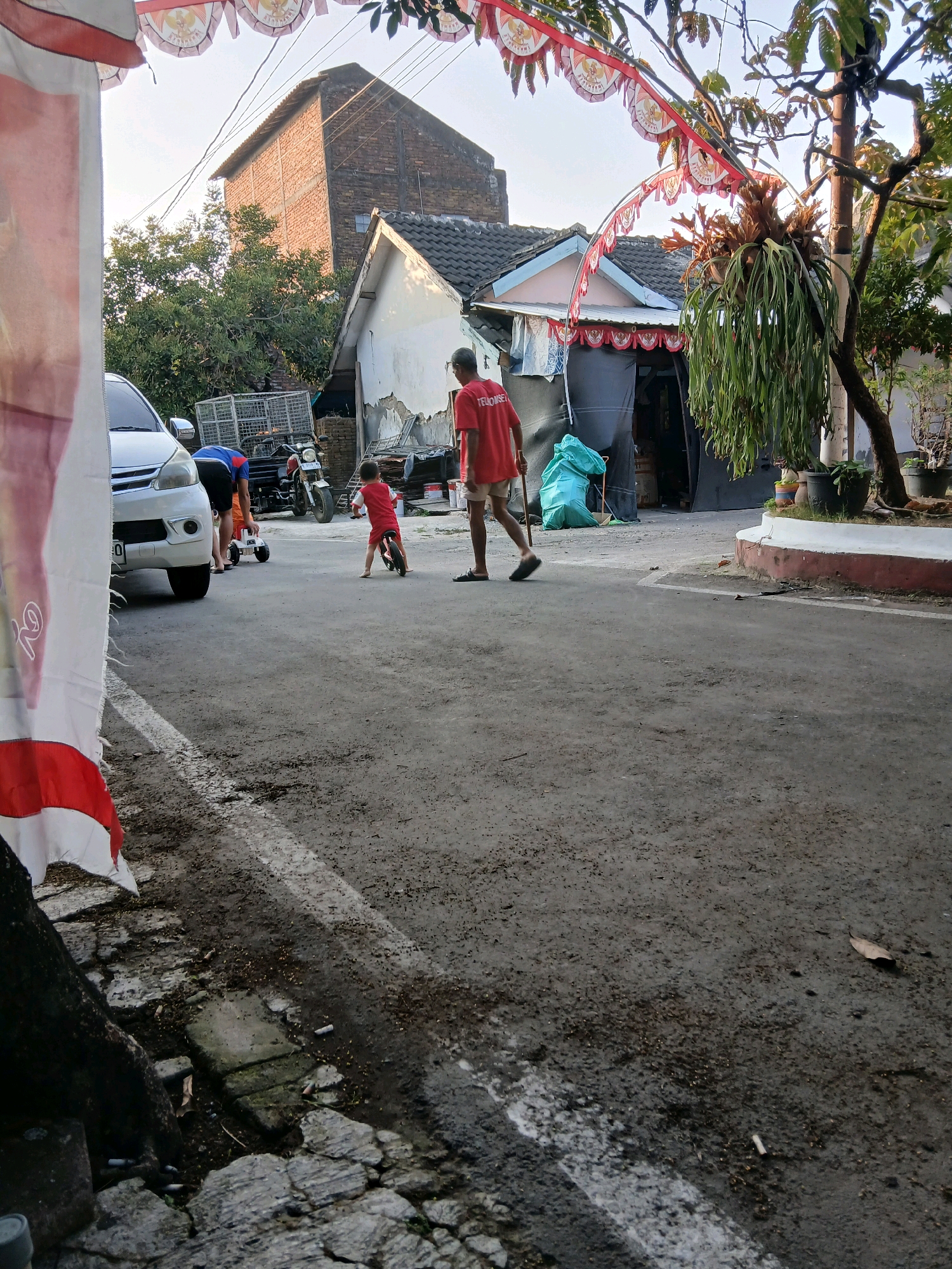 In Jepangpakis, Indonesia, a street scene unfolds with a focus on two individuals and a child. A man in a red t-shirt and light-colored shorts stands on the right side of the asphalt road, facing away from the camera and holding a stick. To his left, a child in a red outfit is on a small bicycle. Further to the left, a white car is parked. Behind these figures, a motorcycle is visible, as well as a large brick building and a smaller house. The environment suggests a residential area with trees and overhead wires. Decorations, possibly flags or banners, are strung across the upper part of the image.