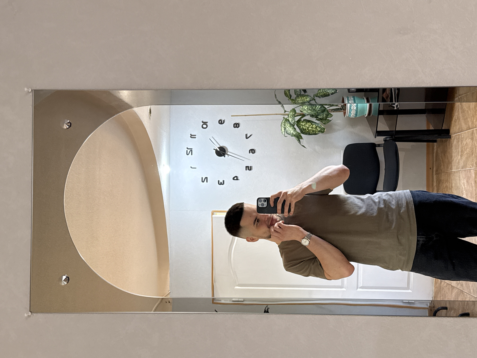 A man is taking a mirror selfie in what appears to be a room in Vereshchagino, Russia. He is wearing a short-sleeved olive green t-shirt and dark pants. On his left wrist, he wears a silver watch. He is holding a smartphone in his right hand, also used to take the photo. His left hand is near his face, as if adjusting his beard or chin. Reflected in the mirror, behind him, is a white door, a black office chair, a small black table with a potted plant, and a wall clock. The room has beige tiled floors and a light-colored wall. The ceiling is visible in the reflection and features a curved architectural element with lights. The overall impression is of a domestic or personal space.