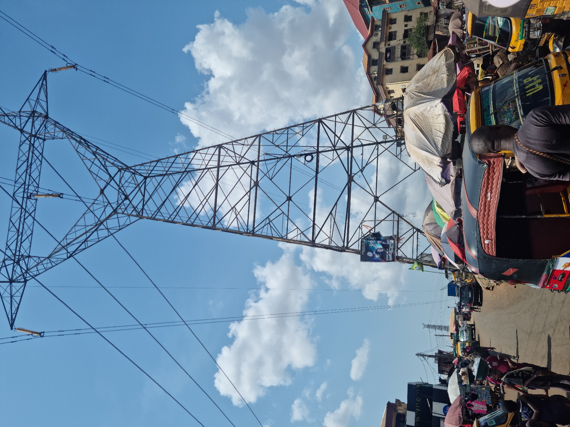 The image captures a vibrant daytime street scene in Obosi, Nigeria, viewed from a slightly elevated or eye-level perspective looking down the street. The sky is a clear blue with scattered white, fluffy clouds.
A prominent feature is a colossal metal electricity transmission tower, or pylon, which occupies a significant portion of the left and center-left of the frame. Its intricate lattice structure is silhouetted against the bright sky, and multiple power lines extend from it across the upper part of the image.
Below the pylon, the scene unfolds into a bustling urban landscape. A street stretches into the distance, teeming with activity. It is lined with numerous market stalls covered by white and colorful tarpaulins and umbrellas, suggesting an active marketplace. Many people are visible on the street, some walking, others congregating around the stalls, engaging in daily commerce.
Several yellow tricycles, locally known as "keke Napeps," are present, parked along the roadside and possibly moving. One is prominently featured in the mid-ground on the right, and another partially visible in the immediate foreground. In the background, multi-story buildings, some with red roofs, indicate a developed residential or commercial area.
Attached to the upper structure of the pylon, a large billboard is visible. It displays an image of several people and some discernible text, including "STREAM OF LIVE FOR" and possibly "YOUNG" or "YOUTH" and "FUTURE." The bright sunlight indicates it is daytime. The image portrays the energetic daily life of an African market town, with essential infrastructure integrated into a lively community setting.