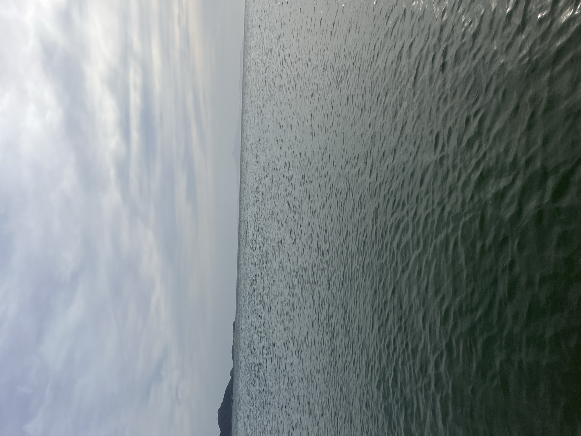 The image shows a body of water, likely the sea, with small waves rippling across its surface. The water is a dark grayish-green color. In the background, there is a sliver of land with a dark, mountainous or hilly silhouette, with a cloudy sky above. The location is likely Juan Griego, Venezuela, given the context. There are no people visible in the image. The dominant subjects are the water and the sky. The interaction is between the wind and the water creating the waves. The environment is overcast and coastal.