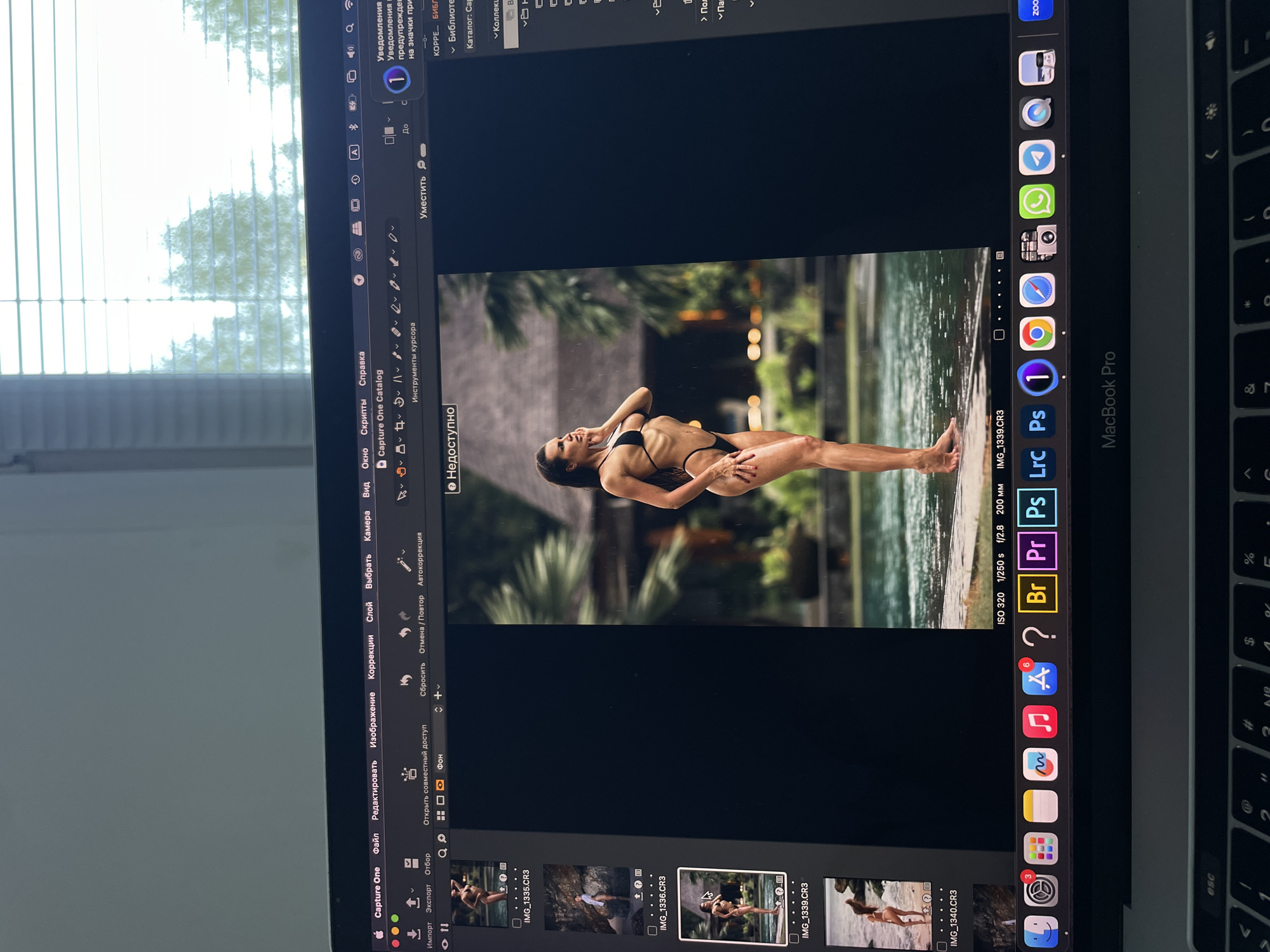 The image shows a laptop screen displaying Capture One software with a photograph of a woman in a black bikini. The woman is positioned horizontally in the image, possibly in or near a body of water with tropical vegetation in the background. The Capture One software interface is visible with various editing tools and file thumbnails. The laptop is a MacBook Pro. The laptop is near a window with the blinds closed. The geographical location of Kazan, Russia is not discernible from the image.