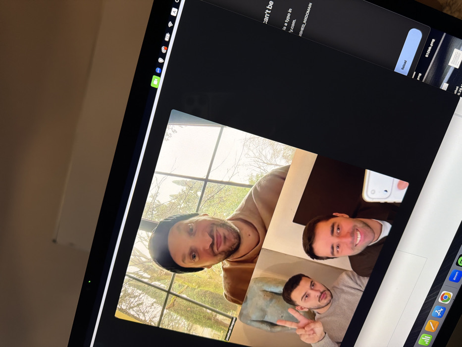 The scene depicts a laptop screen displaying a video conference and a partially visible web browser interface, set within the city of Tel-Aviv, Israel. The left portion of the screen features a video call with three male individuals. In the largest, upper frame, a man with a beard and black beanie smiles at the camera, wearing a light brown shirt. He is identified by a partially visible name as 'Jonathan Saragossi'. Behind him, a large multi-paned window reveals an outdoor environment with numerous bare trees and bright, natural light. In the lower left section of the video call, two other men are visible in separate frames. The man on the left, wearing a grey sweater, makes a peace sign with his right hand; a light blue/green torso sculpture is partially visible next to him. The man on the right smiles while holding a white smartphone, appearing to take a selfie or record himself. The right side of the laptop screen displays a dark-themed web browser with an error message ("can't be", "SHED_NXDOMAIN") and a "Reload" button, alongside partial file names like "512bb.png". The laptop's top menu bar shows an active green video camera icon, and the bottom dock displays application icons, with Zoom highlighted, indicating its use for the video call.