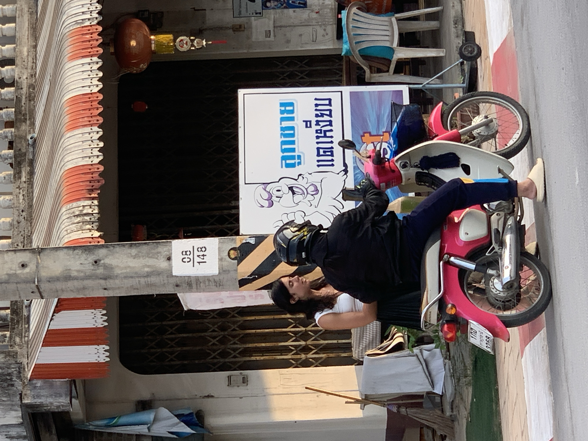 In the city of Ko Samui District, Thailand, a woman wearing a white tank top and a black pleated skirt stands to the left of a motorcycle. She is looking towards the right, in the direction of the motorcycle, where a person in a black jacket and helmet is seated. The motorcycle is pink and white, with a license plate reading "108 3268". A sign with a cartoon chicken and Thai text is visible behind the motorcycle. In the background, a building with a striped awning and an open metal gate is present. The scene takes place on a street with a sidewalk marked with red and white lines.