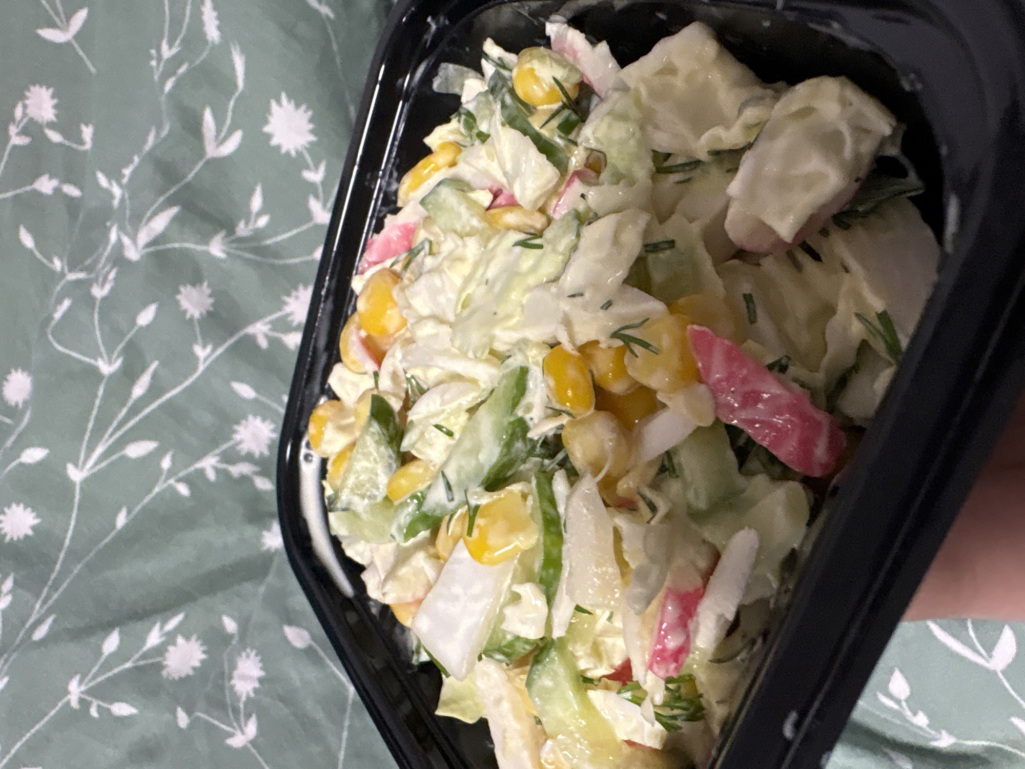 The image shows a black plastic container filled with a salad. The salad appears to contain corn kernels, sliced cucumbers, imitation crab meat (surimi), potatoes and chopped herbs (possibly dill), all coated in a creamy dressing. The container is placed on a light green fabric with a white floral pattern. The location is Прикамский, Russia.