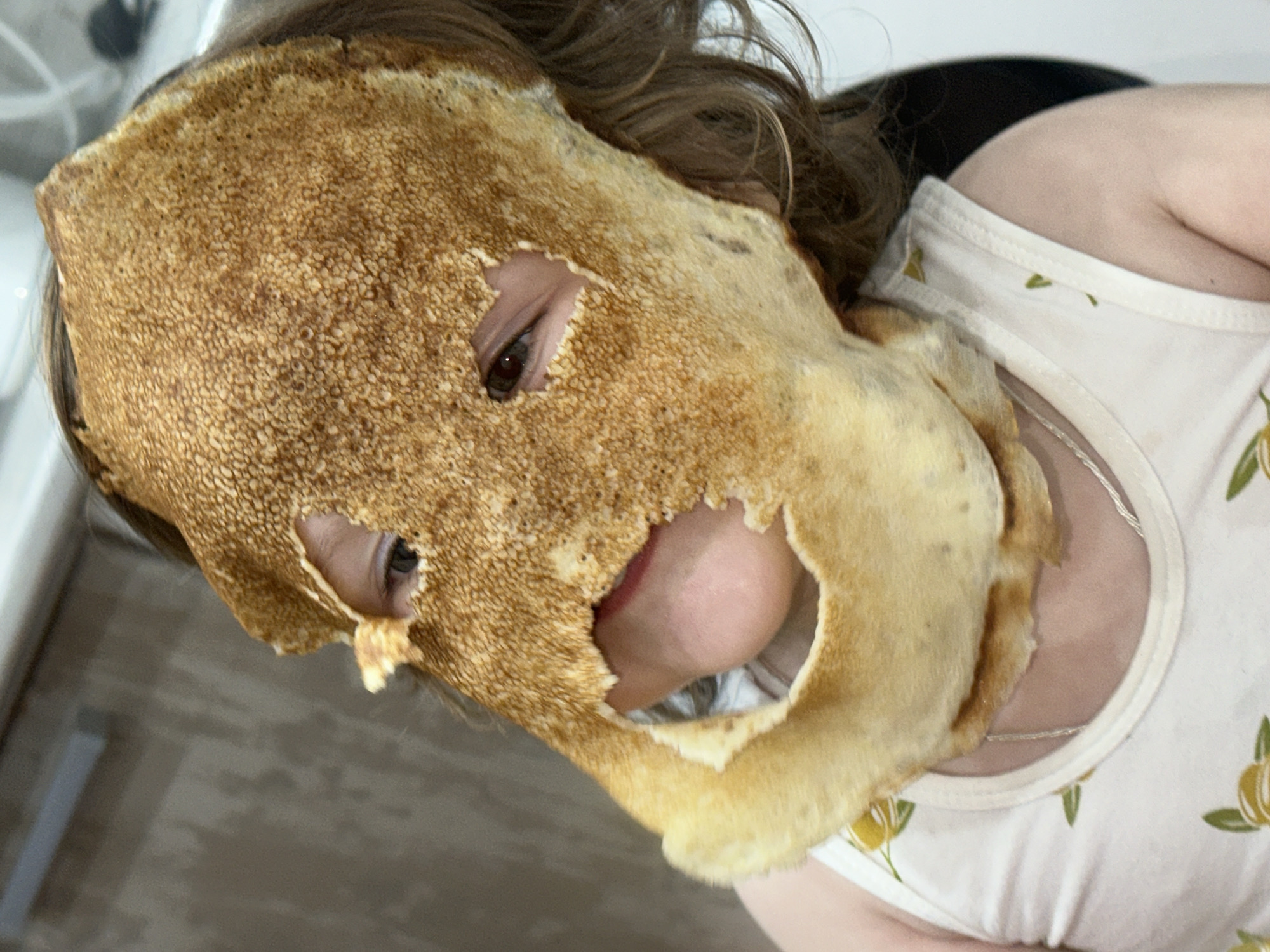 A person is wearing a pancake as a mask. The pancake covers most of their face, with holes cut out for the eyes and mouth. The person has brown hair and is wearing a white shirt with a floral print. The background is blurred, but it appears to be a kitchen or dining area. I have no information about a location in the city of Chaikovsky, Russia, or the city of Chaikovsky itself.
