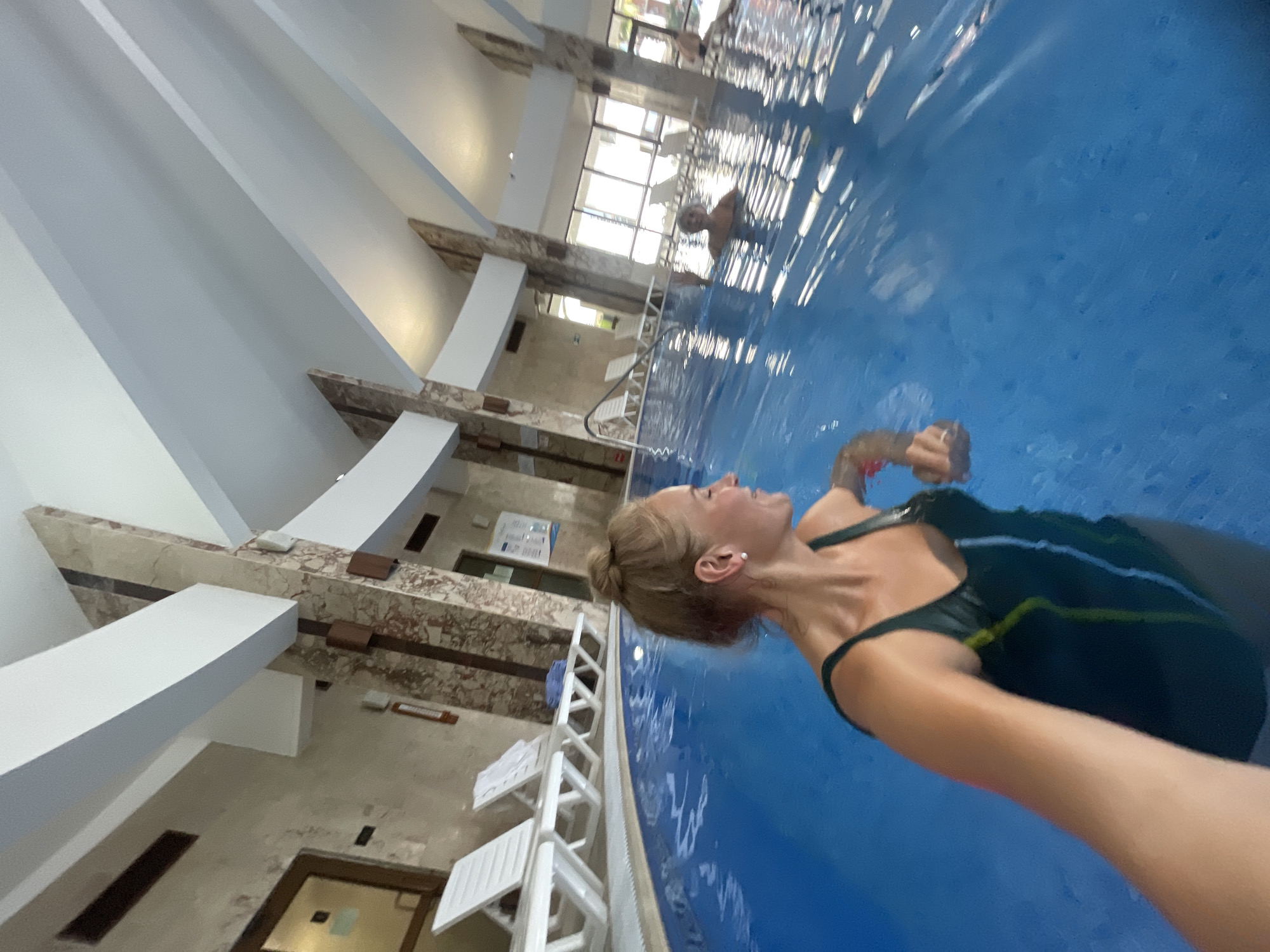 The image shows a person in a swimming pool, likely indoors, with another person visible in the distance. The pool is surrounded by architectural features including white pillars and marble-patterned walls. Outside, there are windows with a view of the sky and potentially other buildings. White chairs are present by the pool. The location is Sochi, Russia.